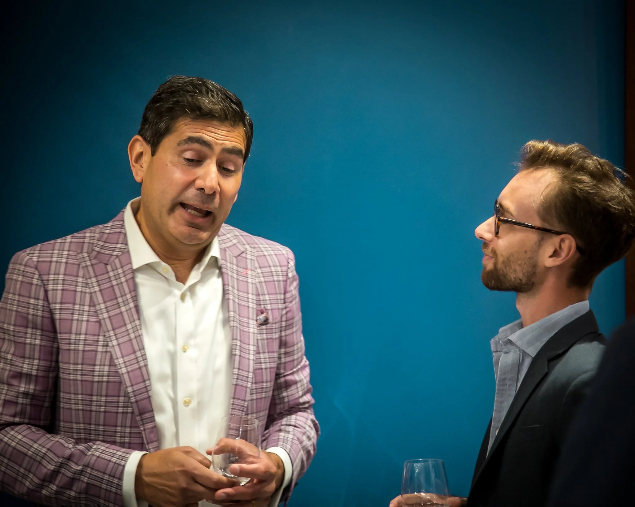 Two men engaged in conversation at a social event, both holding glasses of drink, against a blue wall background.