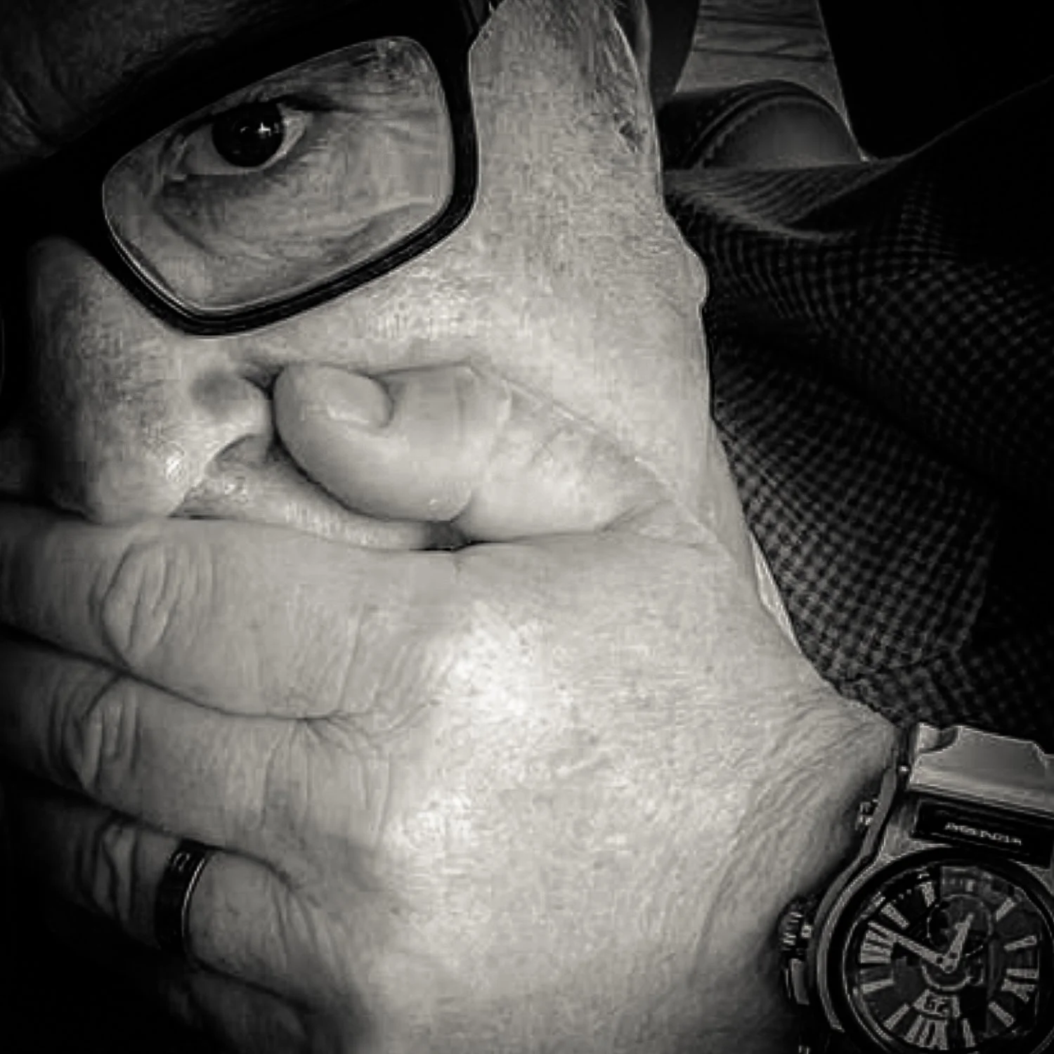Close-up black-and-white photo of a man's face partially covered by his hand, wearing glasses, a watch, and a ring.