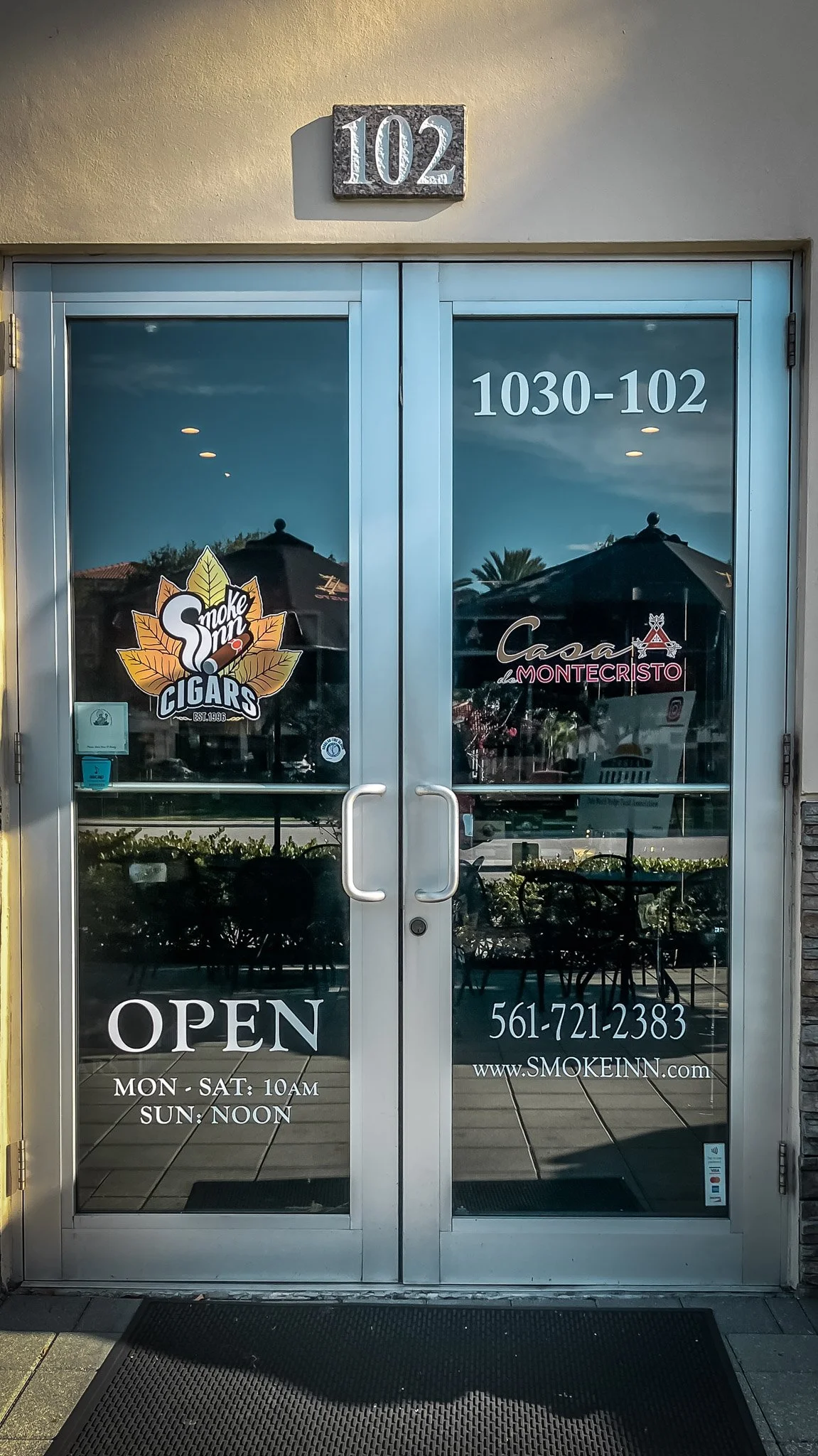 Glass door of a cigar shop with signs for Smoke Inn Cigars and Casa de Montecristo, displaying the address 102, hours, phone number, and website, with reflections of outdoor umbrellas and palm trees.
