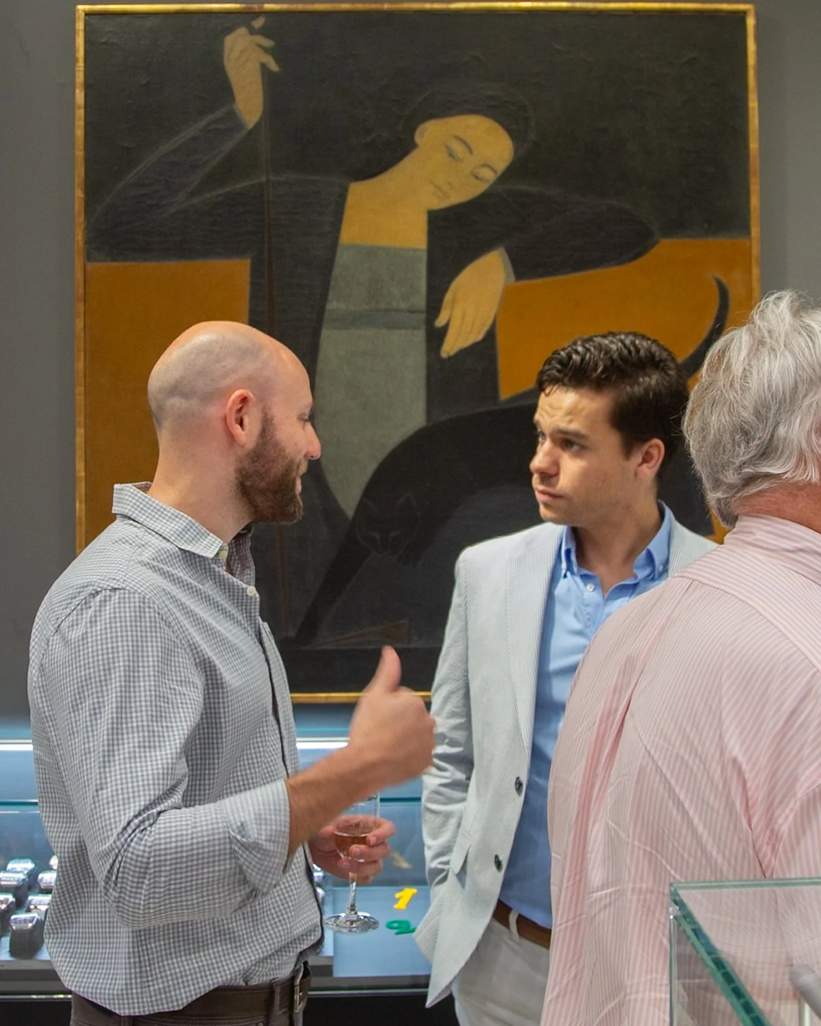 Three men in conversation at a social event, with a modern painting of a woman playing piano in the background.