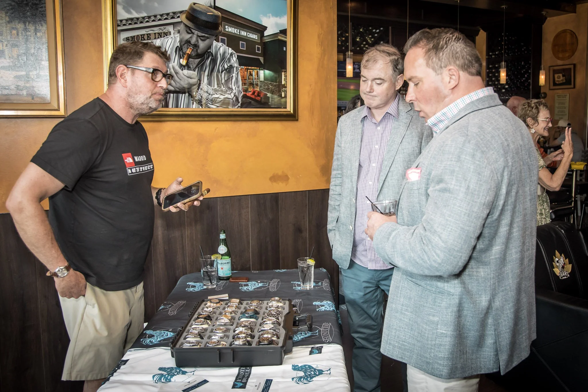 Three men in business casual attire stand at a table looking at a display of watches, with one man holding a smartphone, inside a restaurant or bar with framed art on the wall.