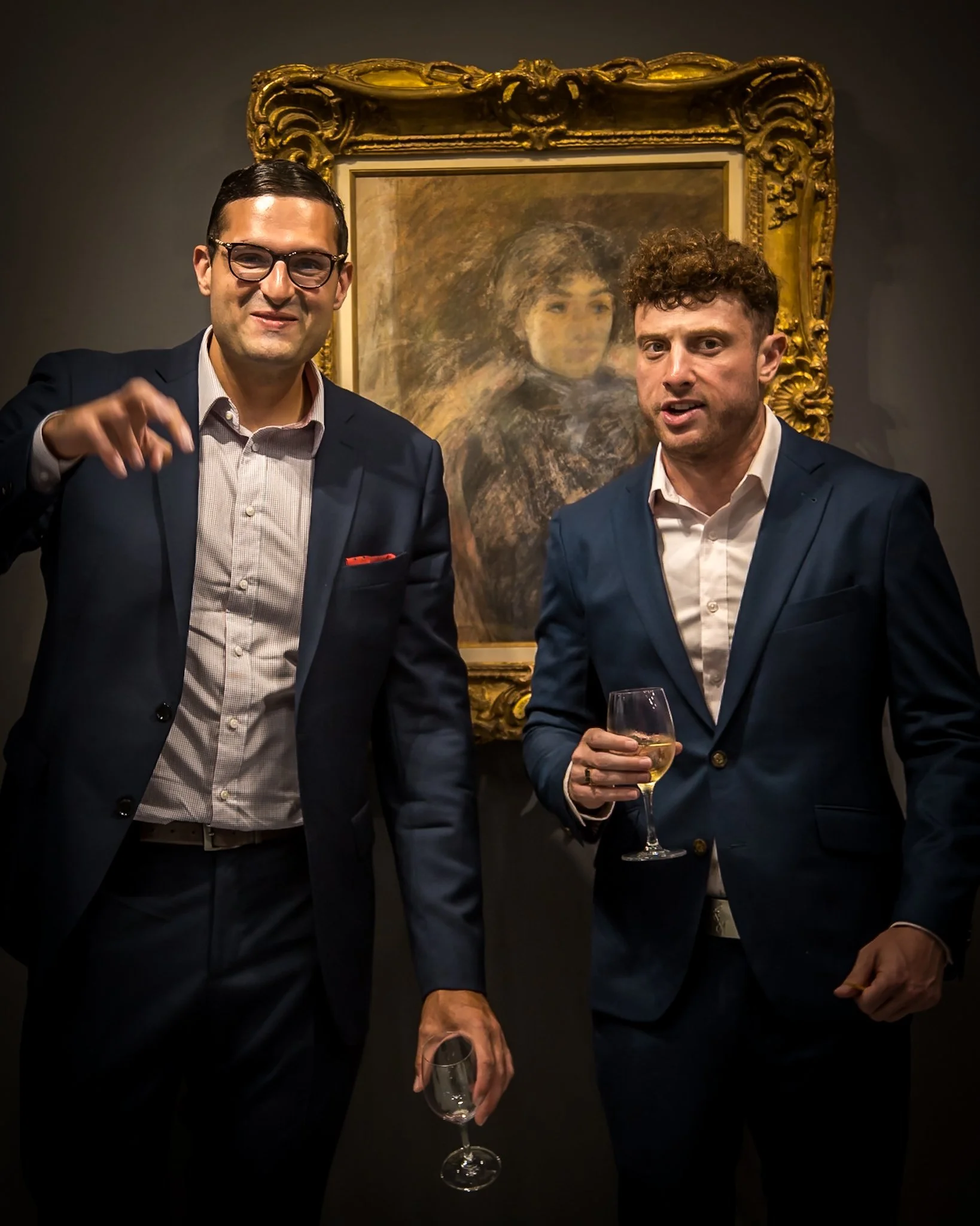 Two men in suits at an art gallery, standing in front of a framed painting, holding glasses of wine, engaging in conversation.