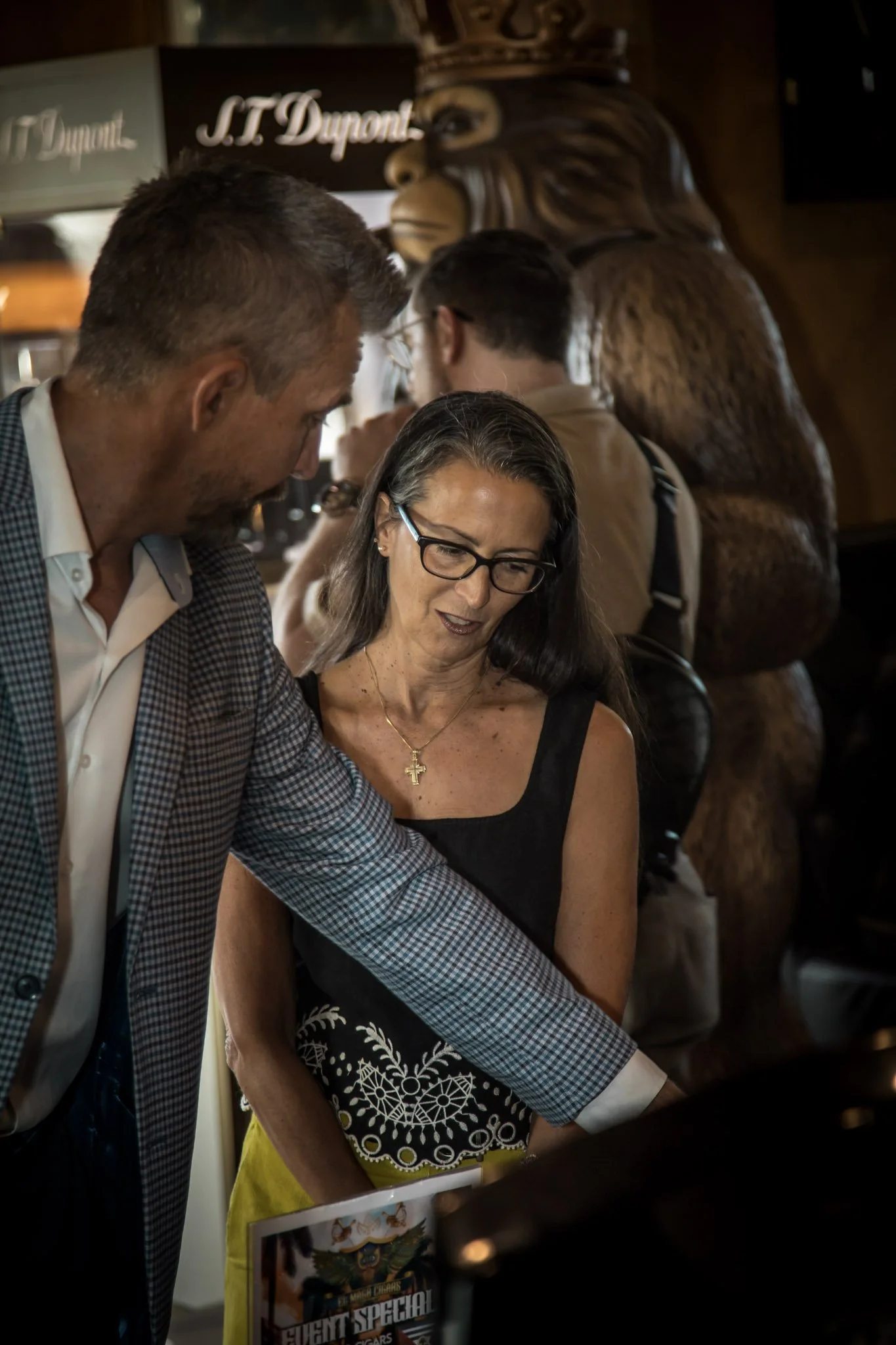 Two people, a man and a woman, are engaged in a conversation at a bar or restaurant. The man is wearing a checkered blazer and white shirt, and the woman is wearing glasses, a black top, and a gold cross necklace. There is a large wooden bear sculptu
