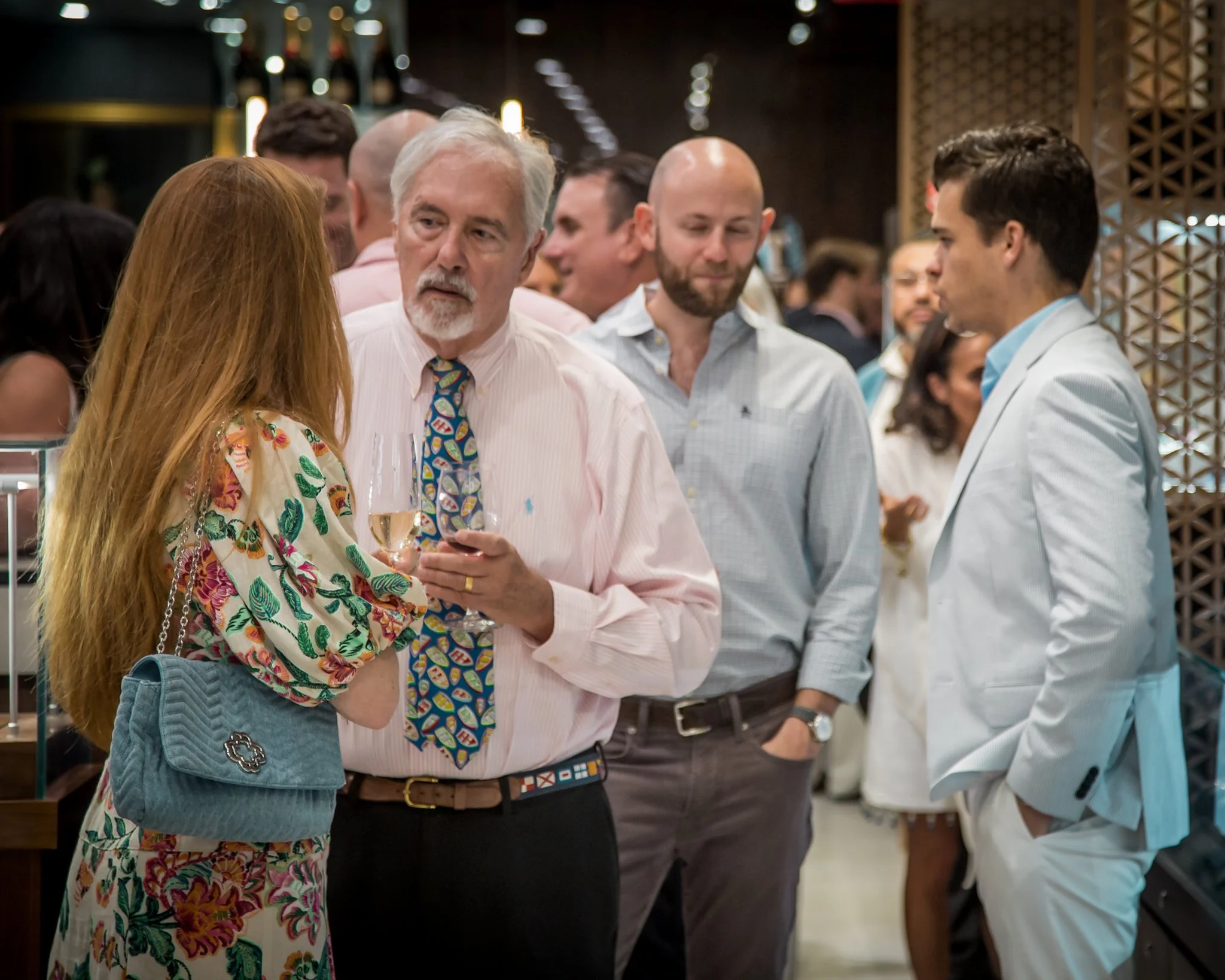A group of people at a social event or party, wearing semi-formal attire, engaged in conversation, with a woman holding a glass of champagne and a man in a pink shirt and colorful tie talking to her.