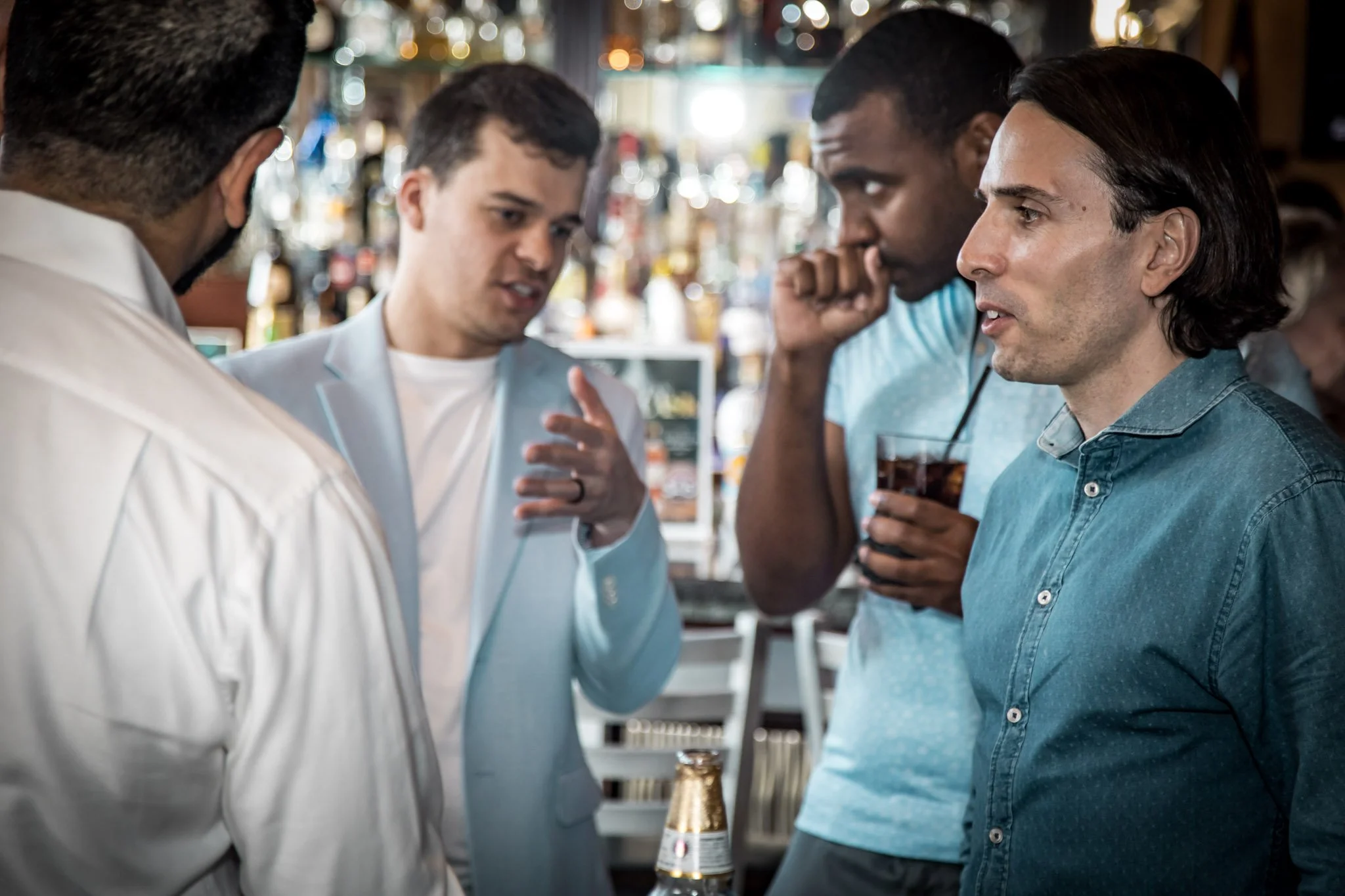Four men engaged in conversation at a bar, one holding a drink.