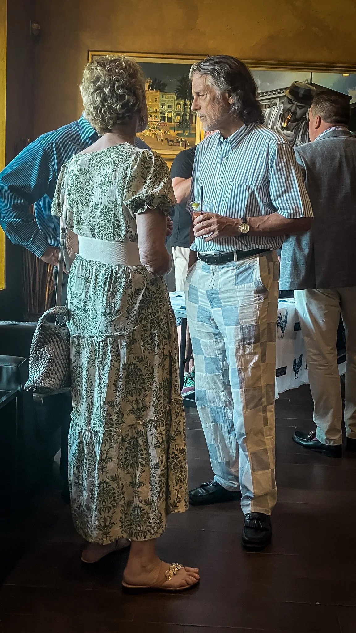 A woman in a patterned dress and sandals converses with a man in a striped shirt and beige checkered pants at a social gathering. Other people are in the background.