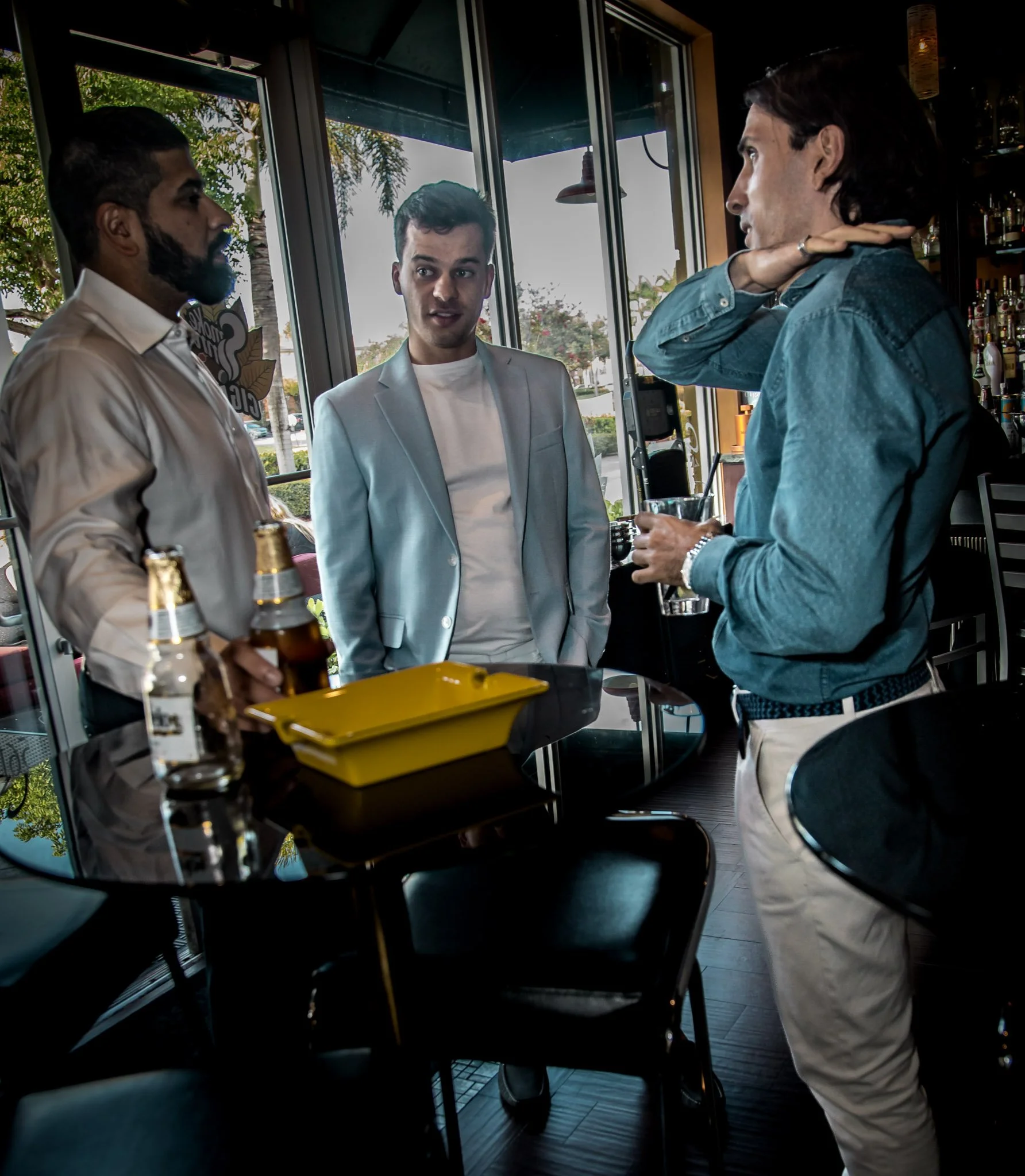 Three men engaged in conversation inside a restaurant or bar, with beverages on the table, large windows revealing trees outside.