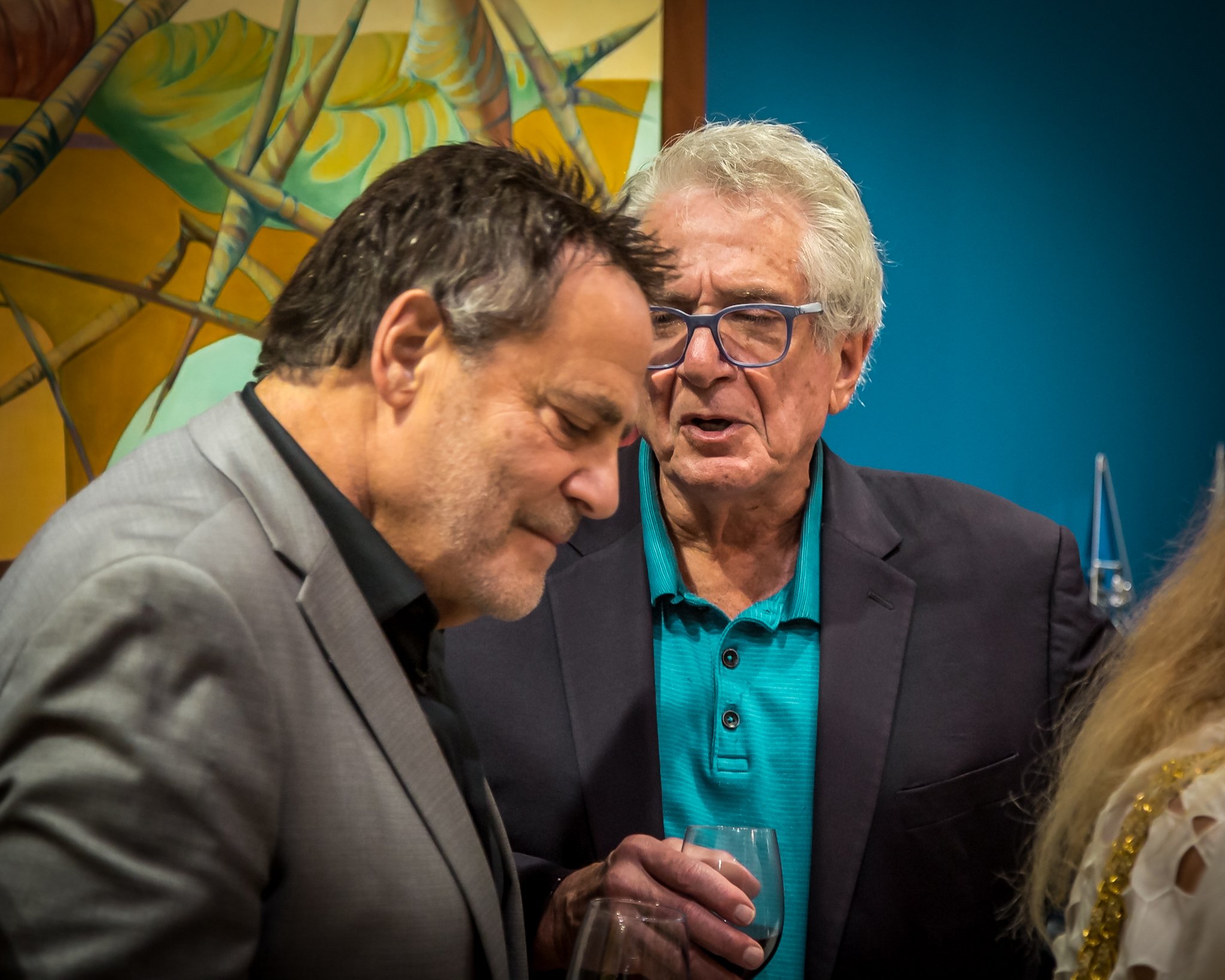 Two men engaged in conversation at a social event, one holding a wine glass, against a background with colorful abstract art.