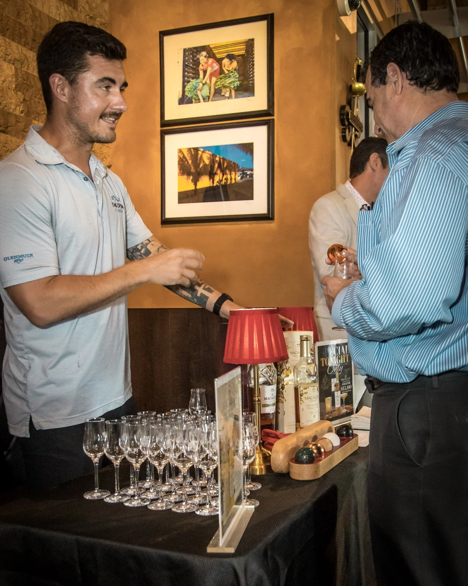 A man with a tattoo on his arm and wearing a gray striped polo shirt is smiling and talking to another man who is holding a glass of rosé wine. They are at a table with empty glasses and bottles of liquor at an indoor event, possibly at a bar or tast