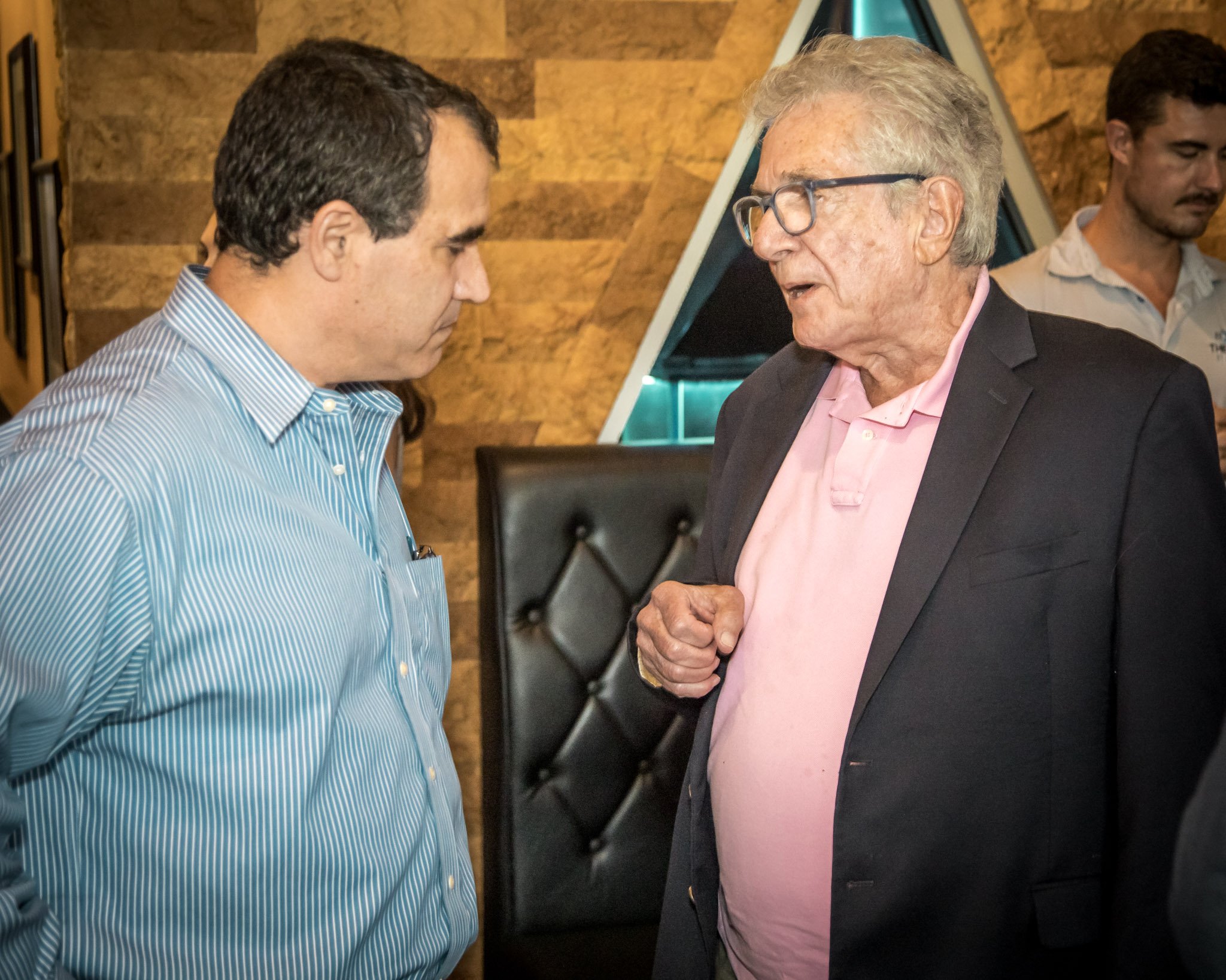 Two men engaged in conversation indoors, with a wooden wall and a decorative background behind them. The man on the left wears a blue and white striped shirt, while the man on the right wears a black blazer over a pink shirt, and glasses.