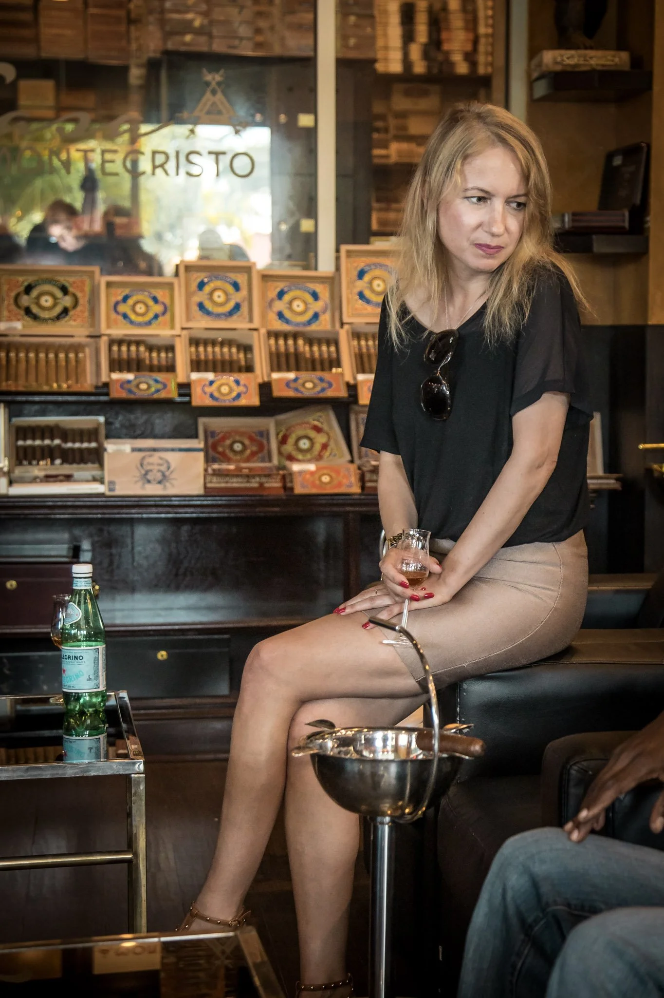 A woman with blonde hair in black attire, sitting on a black sofa holding a glass of champagne, in a cigar lounge with wooden shelves and cigar boxes behind her, and a bottle of San Pellegrino sparkling water on a table in front.