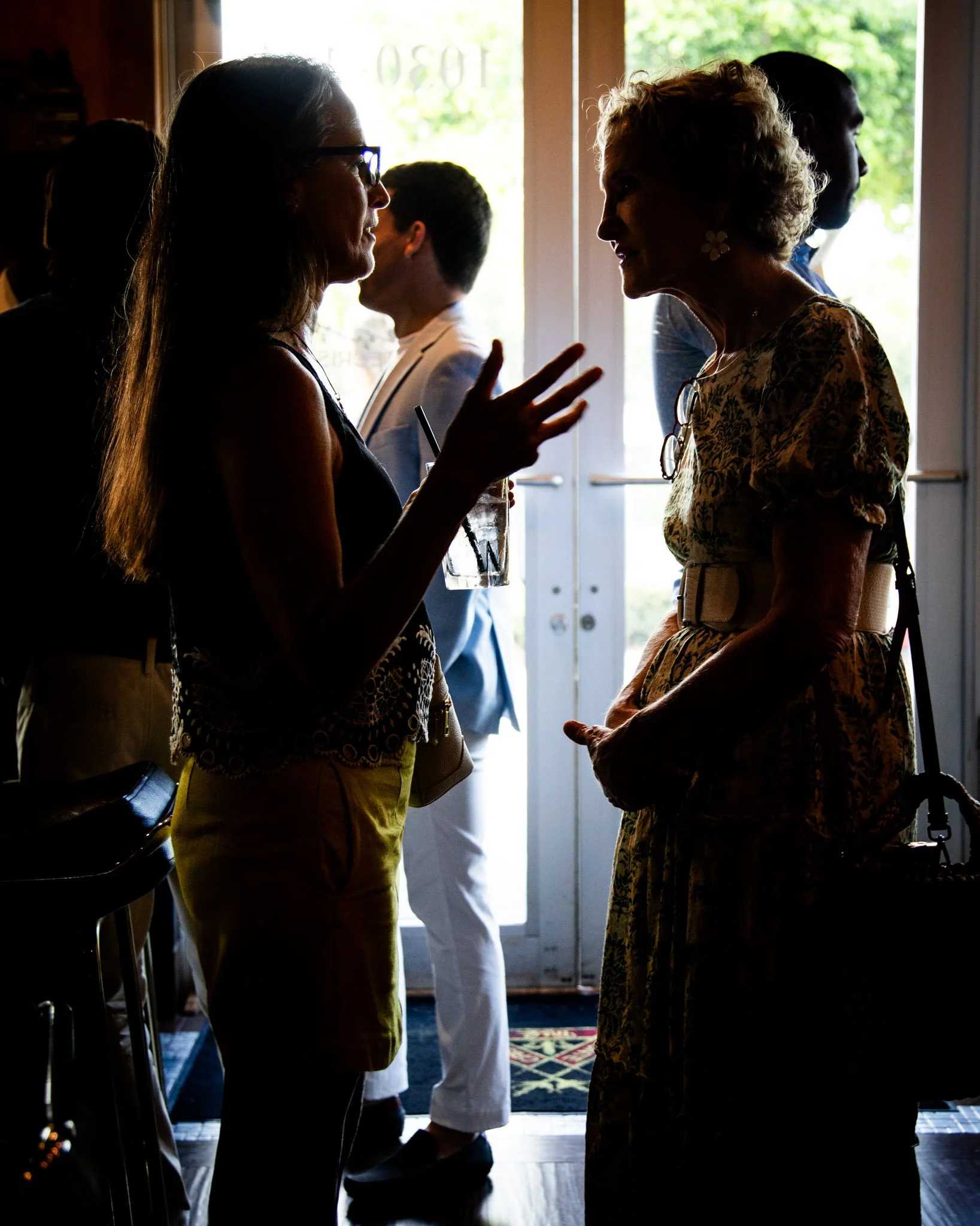 Two women engaged in a conversation in a dimly lit indoor setting with sunlight coming through the windows behind them.