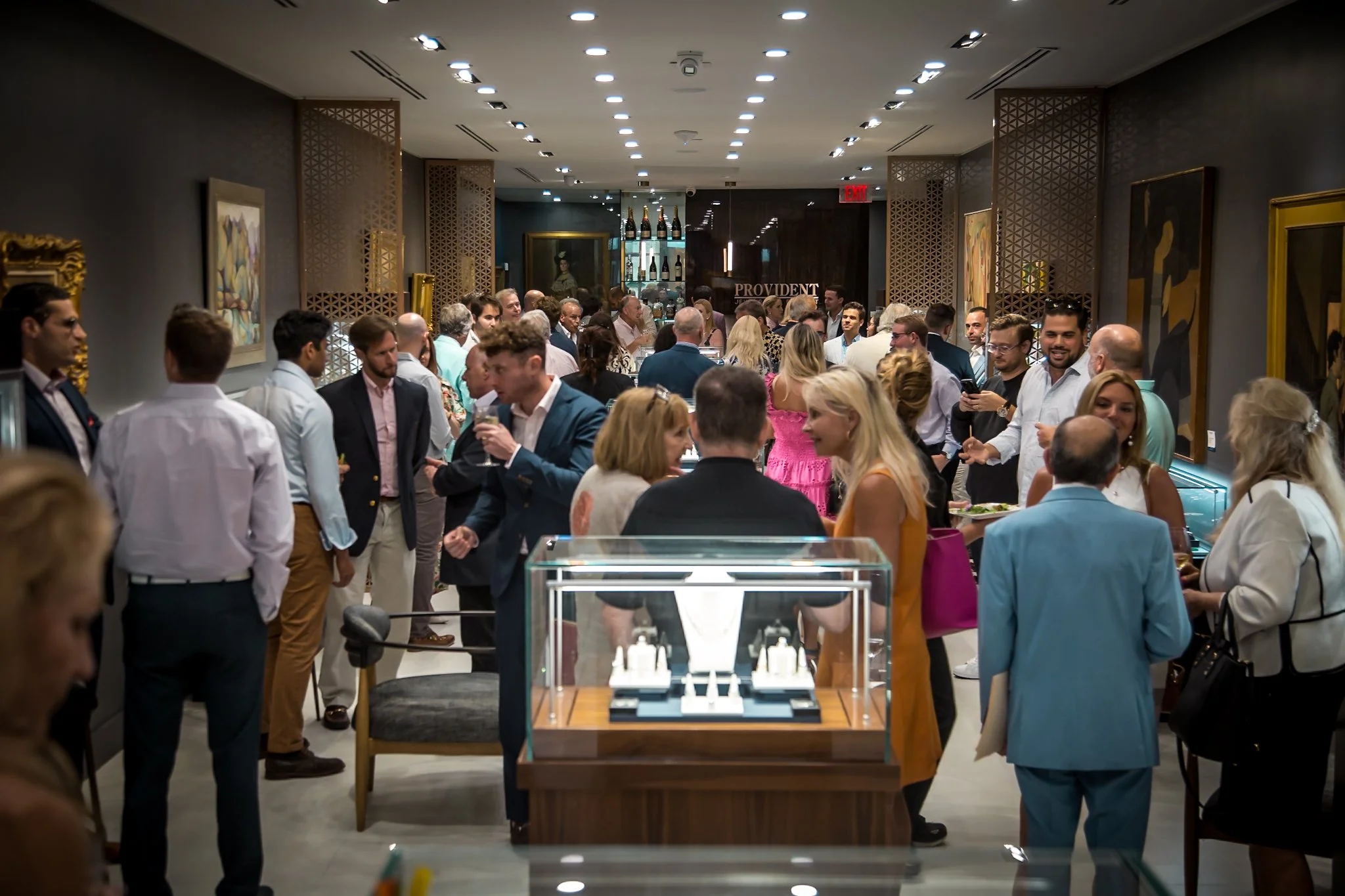 A crowded art gallery or event space with people socializing and looking at artwork on the walls. There are paintings on the walls, display cases with jewelry, and a bar area in the background.