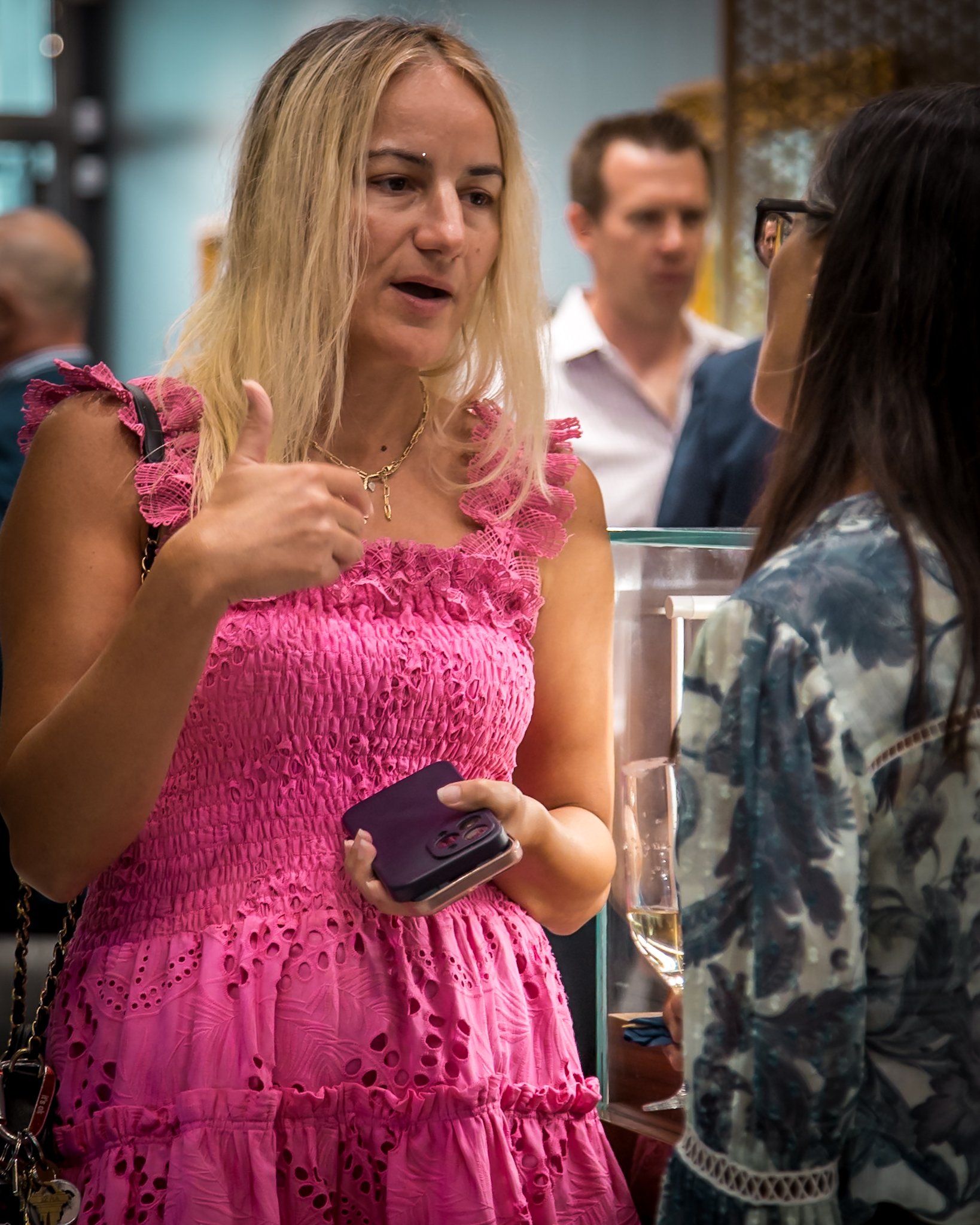 Two women engaged in conversation at an indoor event, one wearing a bright pink dress and holding a smartphone, the other wearing a patterned top.