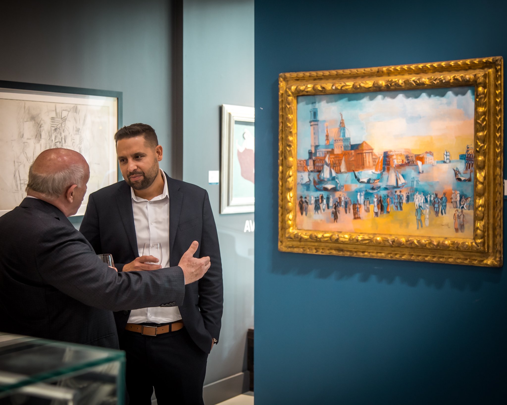 Two men in suits having a conversation and holding glasses near artworks in an art gallery.