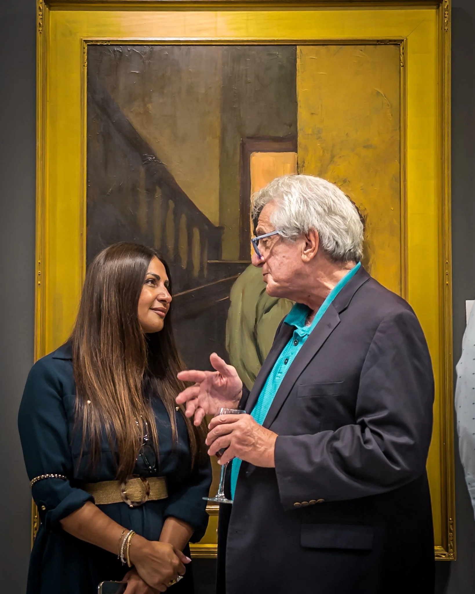 Two people conversing at an art gallery, with a large framed painting of an interior staircase in the background.