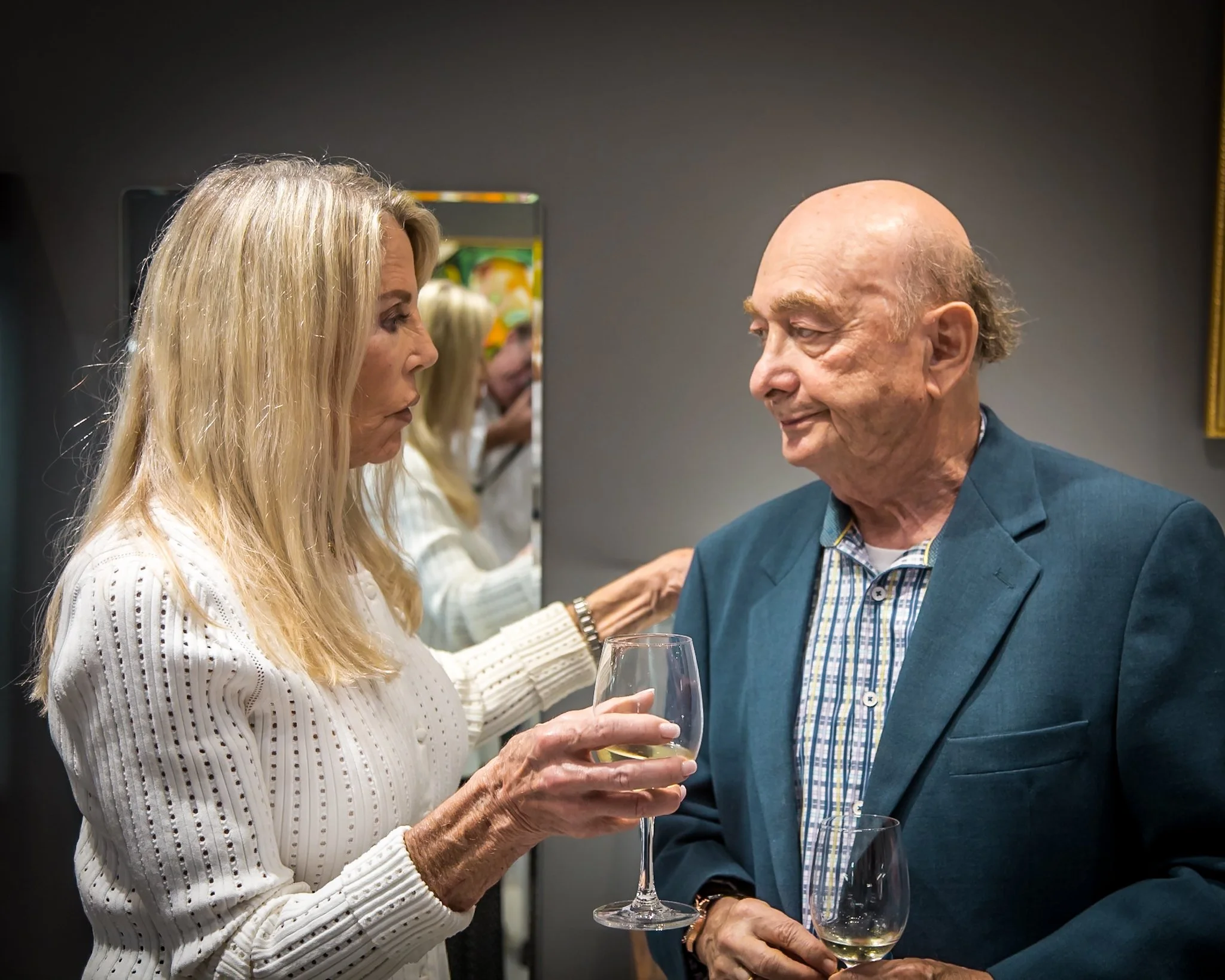 An elderly woman with long blonde hair drinking white wine and talking to an elderly man with a bald head and short hair at a social gathering. The woman wears a white sweater, and the man wears a teal blazer with a checked shirt. They are holding wi