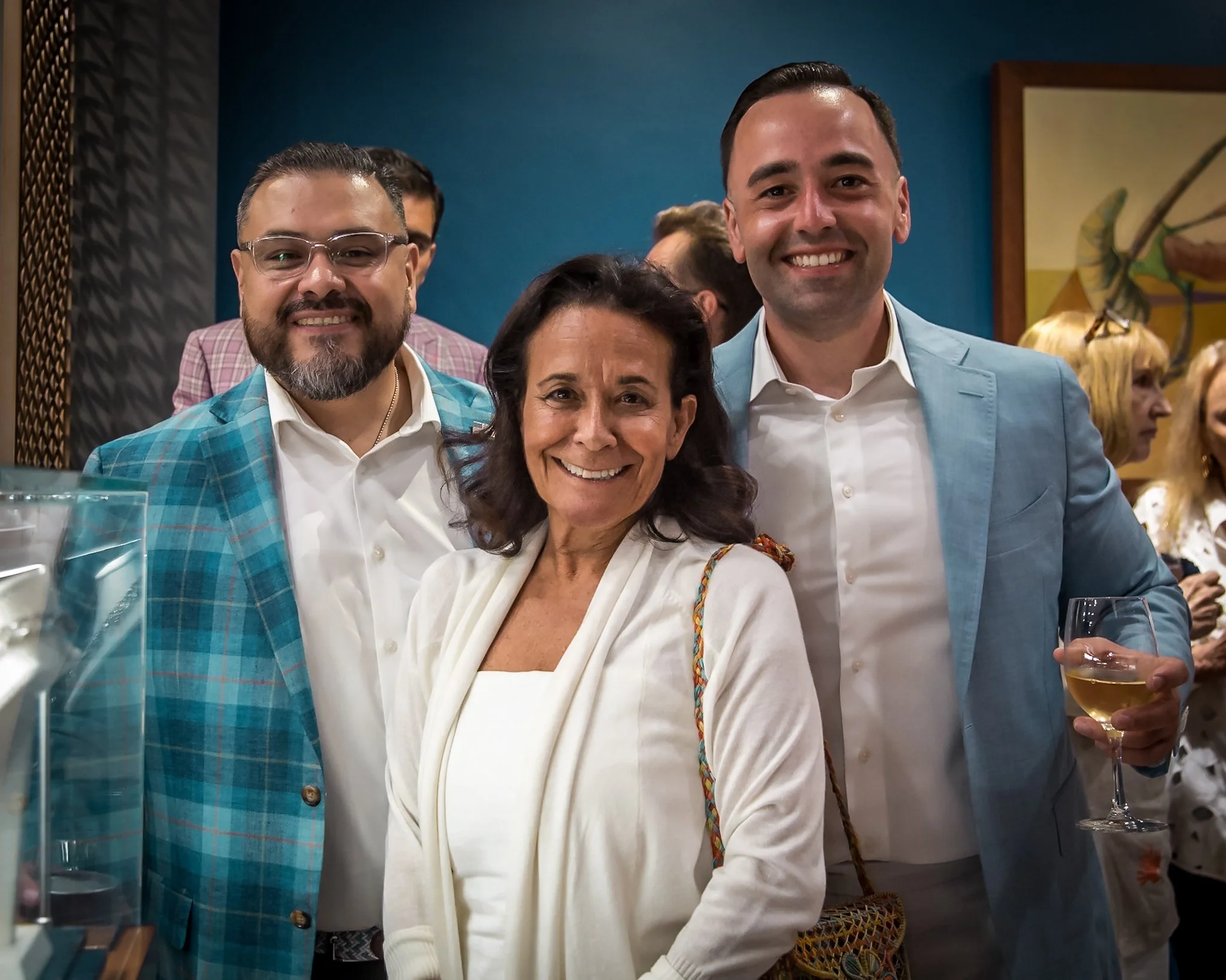 Three people standing closely together at a social event, smiling at the camera. The woman in the middle has dark hair and is wearing a white top, with a patterned bag over her shoulder. The man on the right is wearing a light blue suit jacket and ho