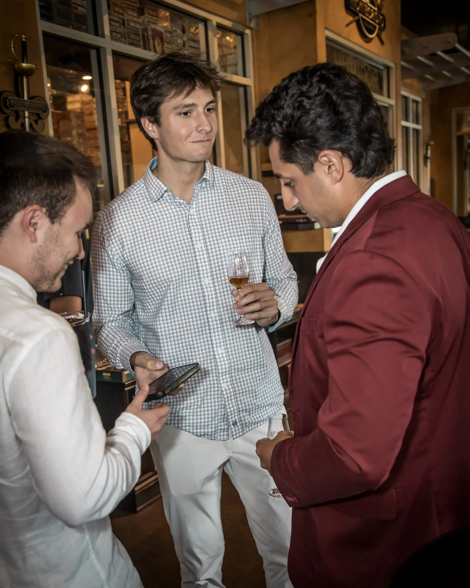 A group of four men socializing indoors, one holding a glass of whiskey, and another looking at a smartphone.