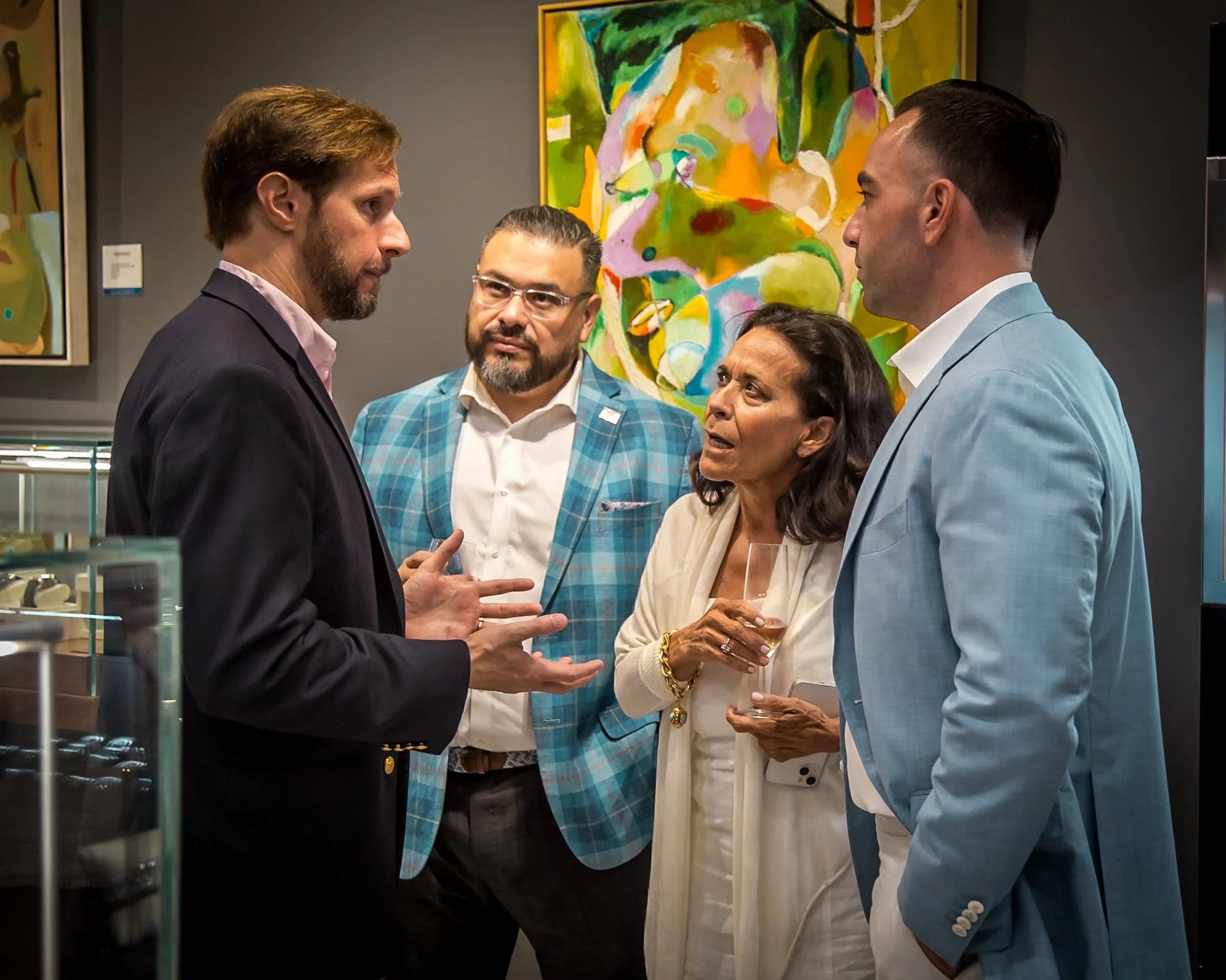Four people engaged in a discussion at an art gallery. A woman with dark hair holding a glass of wine looks concerned, while a man in a black blazer speaks with expressive hand gestures. Two men, one in a turquoise plaid blazer and another in a light