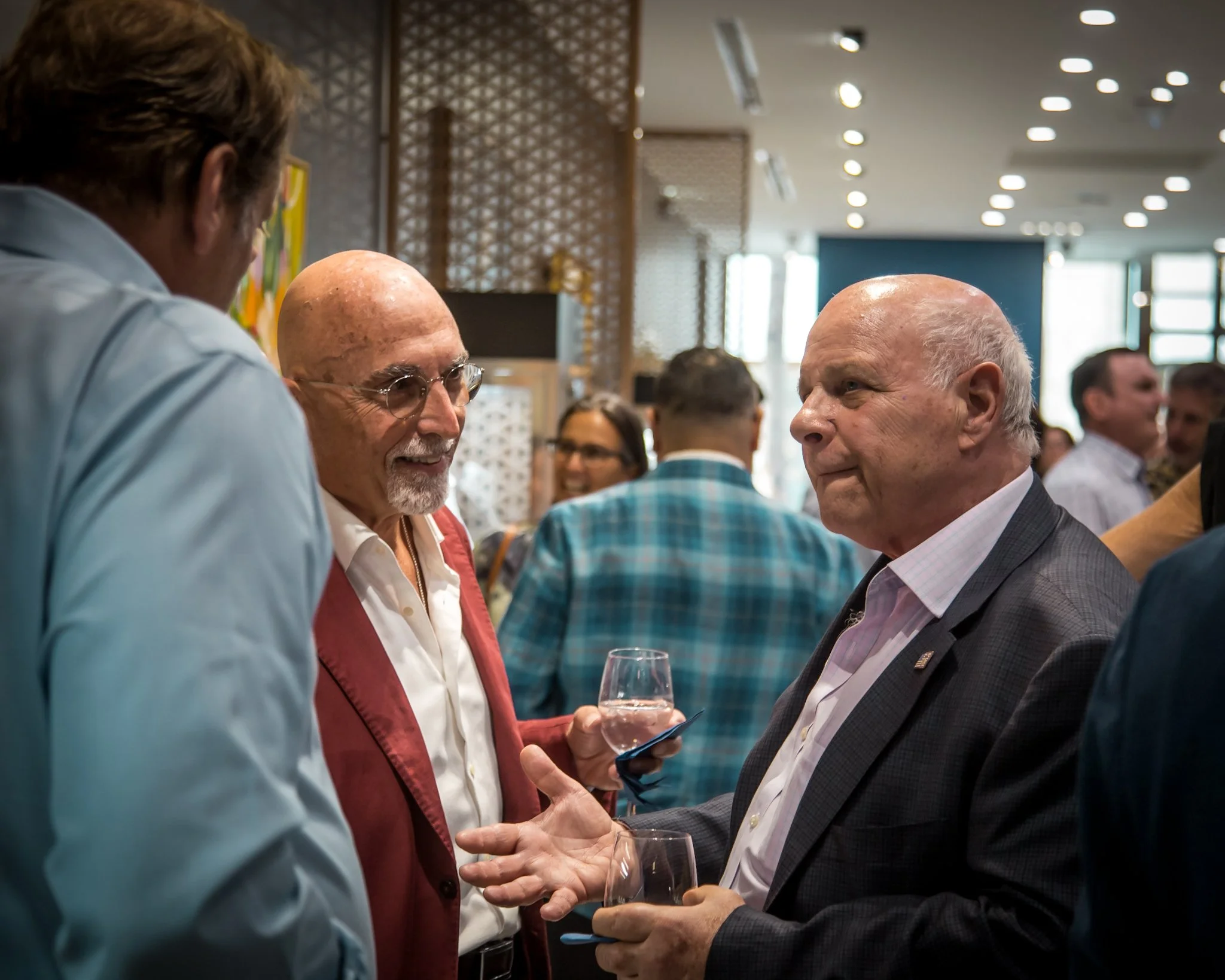 Three men engaged in conversation at an indoor social event, holding drinks, with other people in the background.