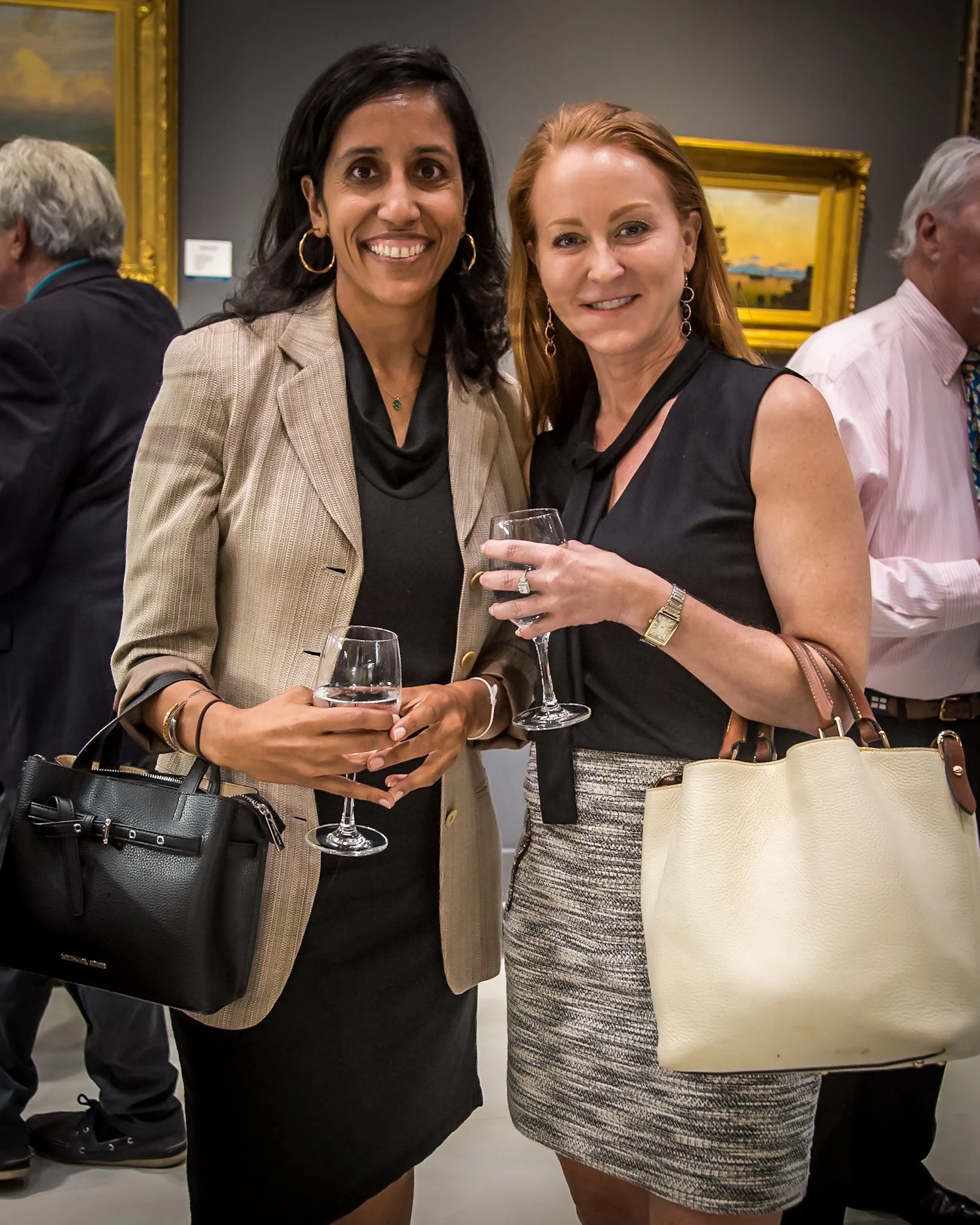 Two women at an art gallery or museum, holding glasses of wine, smiling for the camera, with other people and paintings in the background.