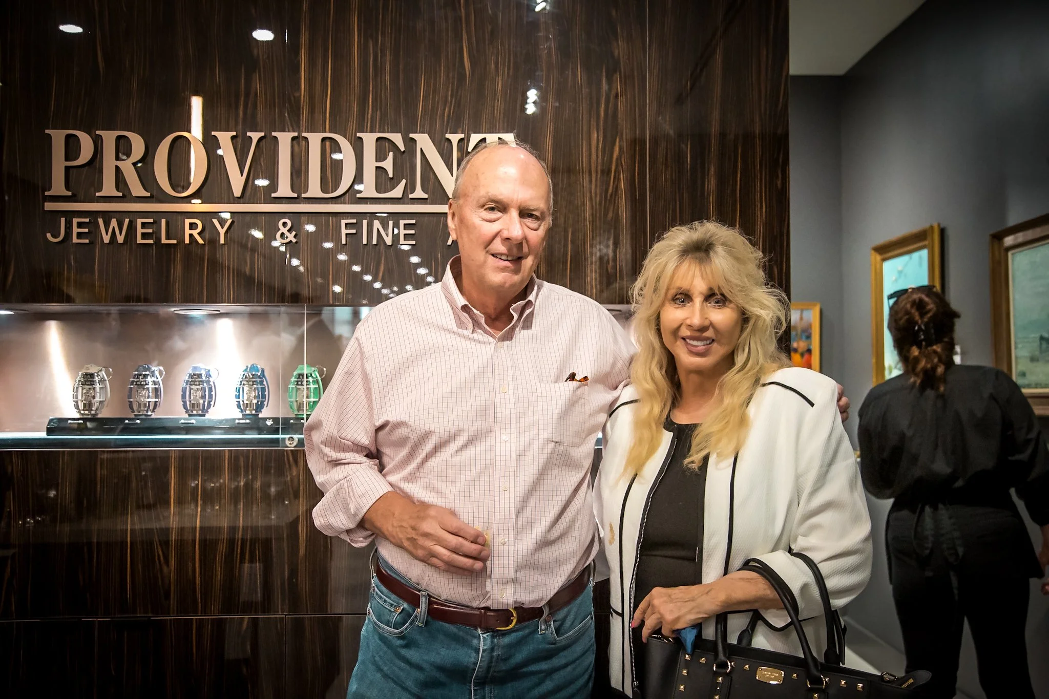 A man and a woman standing together and smiling in front of a jewelry store sign that reads "PROVIDENT JEWELRY & FINE." They are indoors, with additional artwork visible on the walls around them.