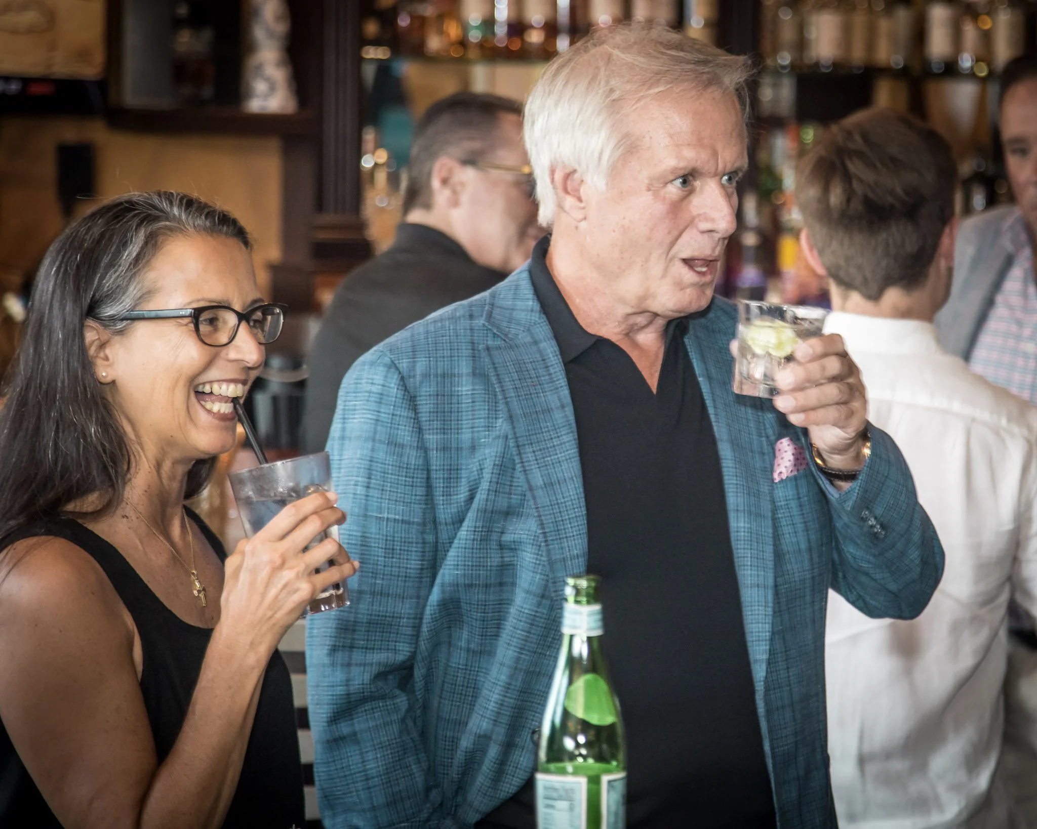 Two people, a woman and a man, are enjoying cocktails at a social gathering. The woman is wearing glasses and a black top, and the man is dressed in a blue checkered blazer and black shirt. They are in a bar or restaurant with other patrons visible i