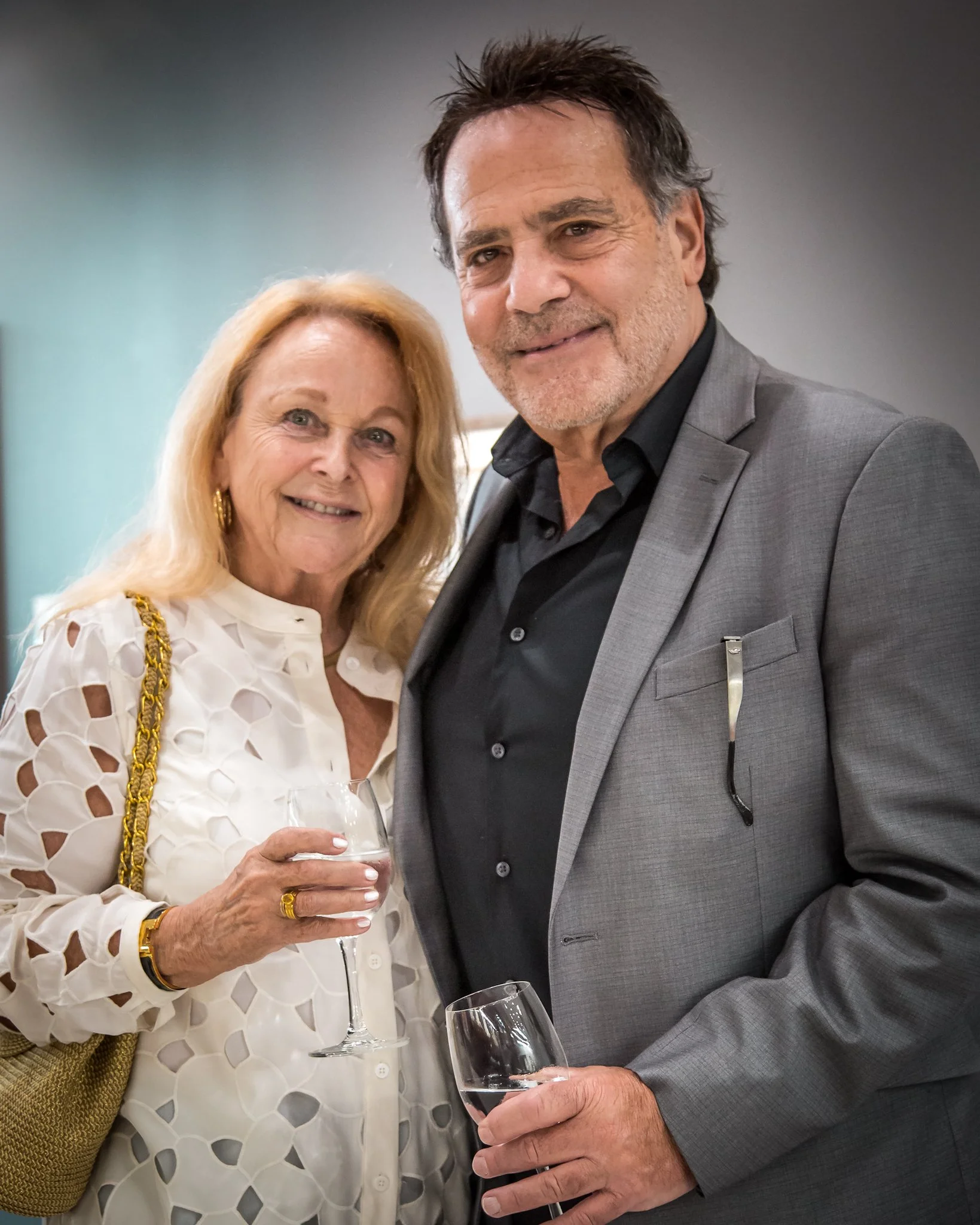 An elderly woman in a white dress holding a wine glass, standing next to a middle-aged man in a gray suit with a black shirt, also holding a wine glass, at a social event.