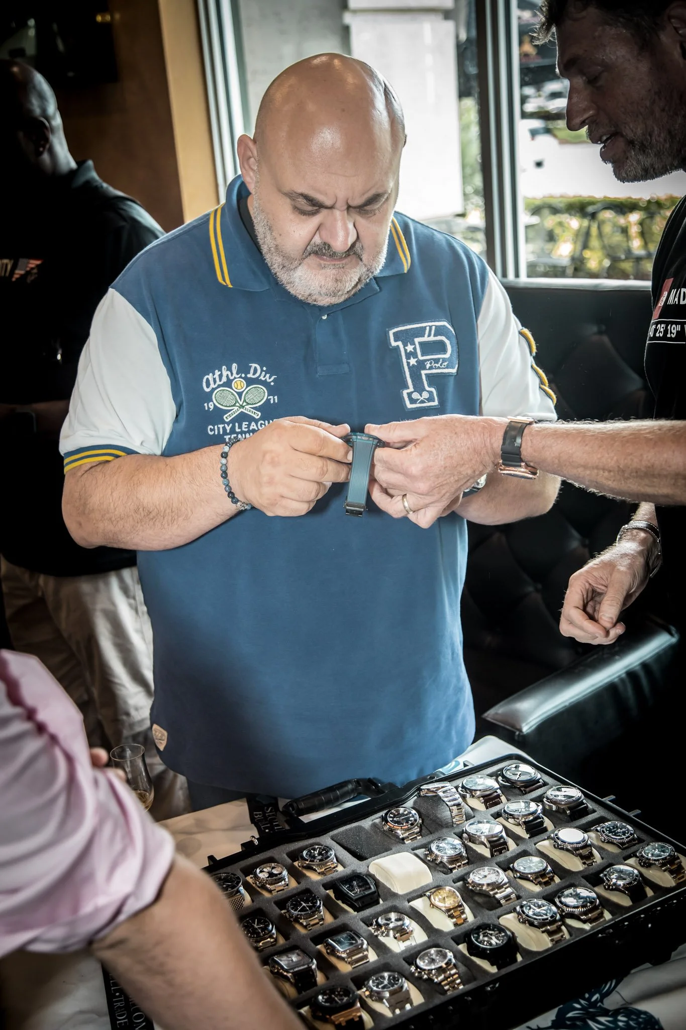 A man is looking at his smartwatch while holding his arm with another person's hand during a watch sale event, with a display case of various watches on the table.