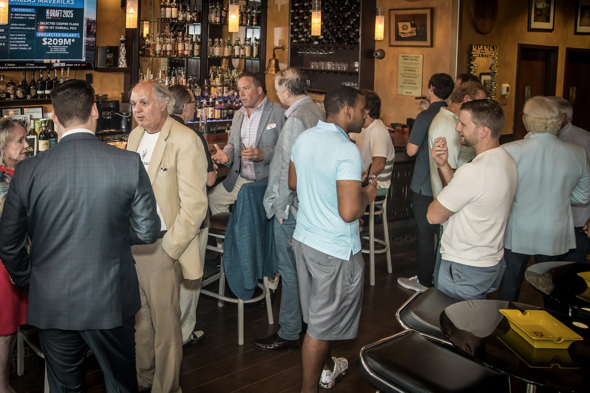 A group of people socializing at a bar or pub, with some standing and some seated, and a bartender serving drinks behind the counter. The bar area has shelves stocked with liquor bottles and a television screen displaying sports news.