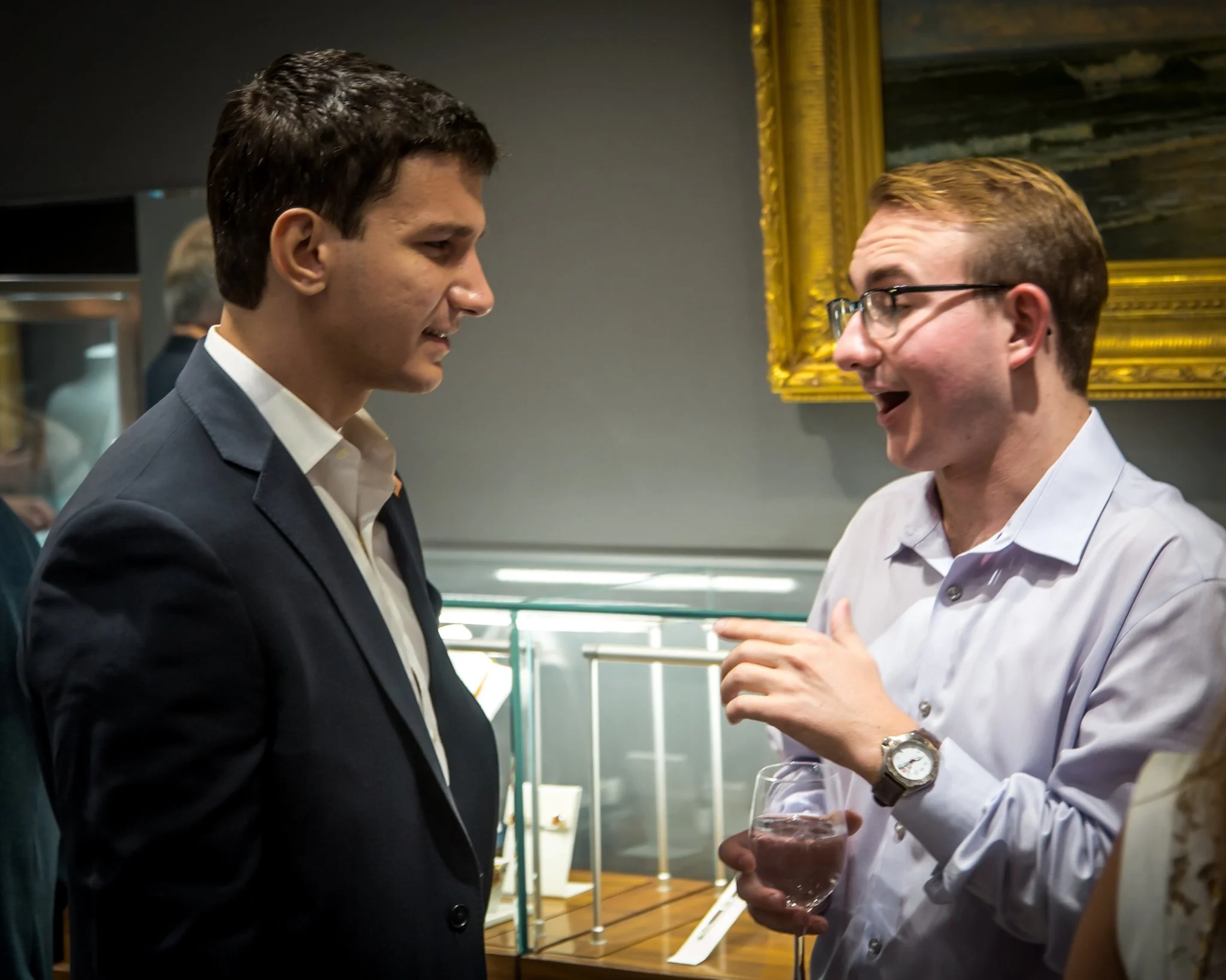 Two men having a conversation at a social event, one in a dark suit and the other in a white shirt with glasses, holding a glass of water, in an indoor setting with artwork and display cases in the background.