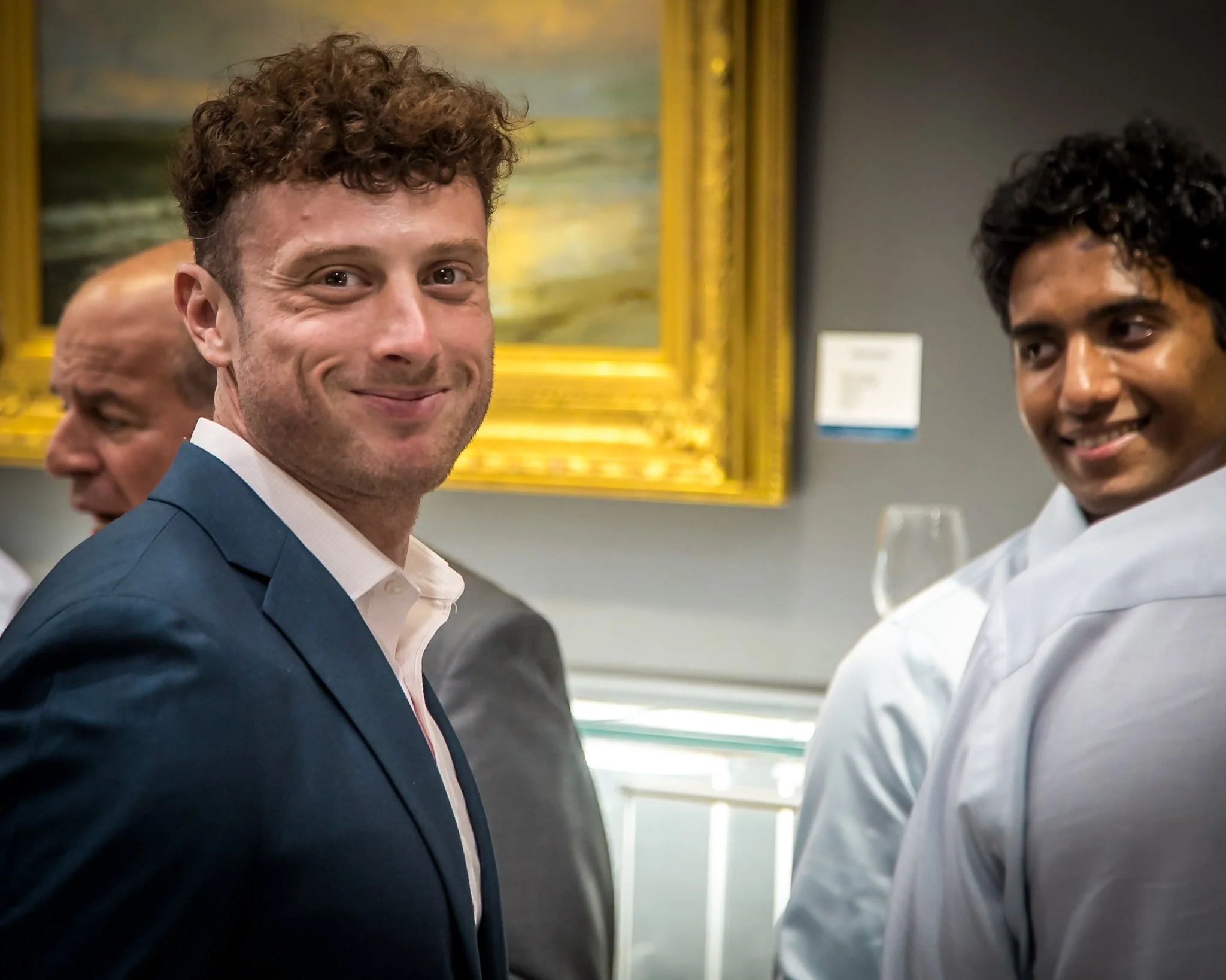 Three men in formal attire at an art gallery, with a painting in a gold frame on the wall behind them, smiling at the camera.