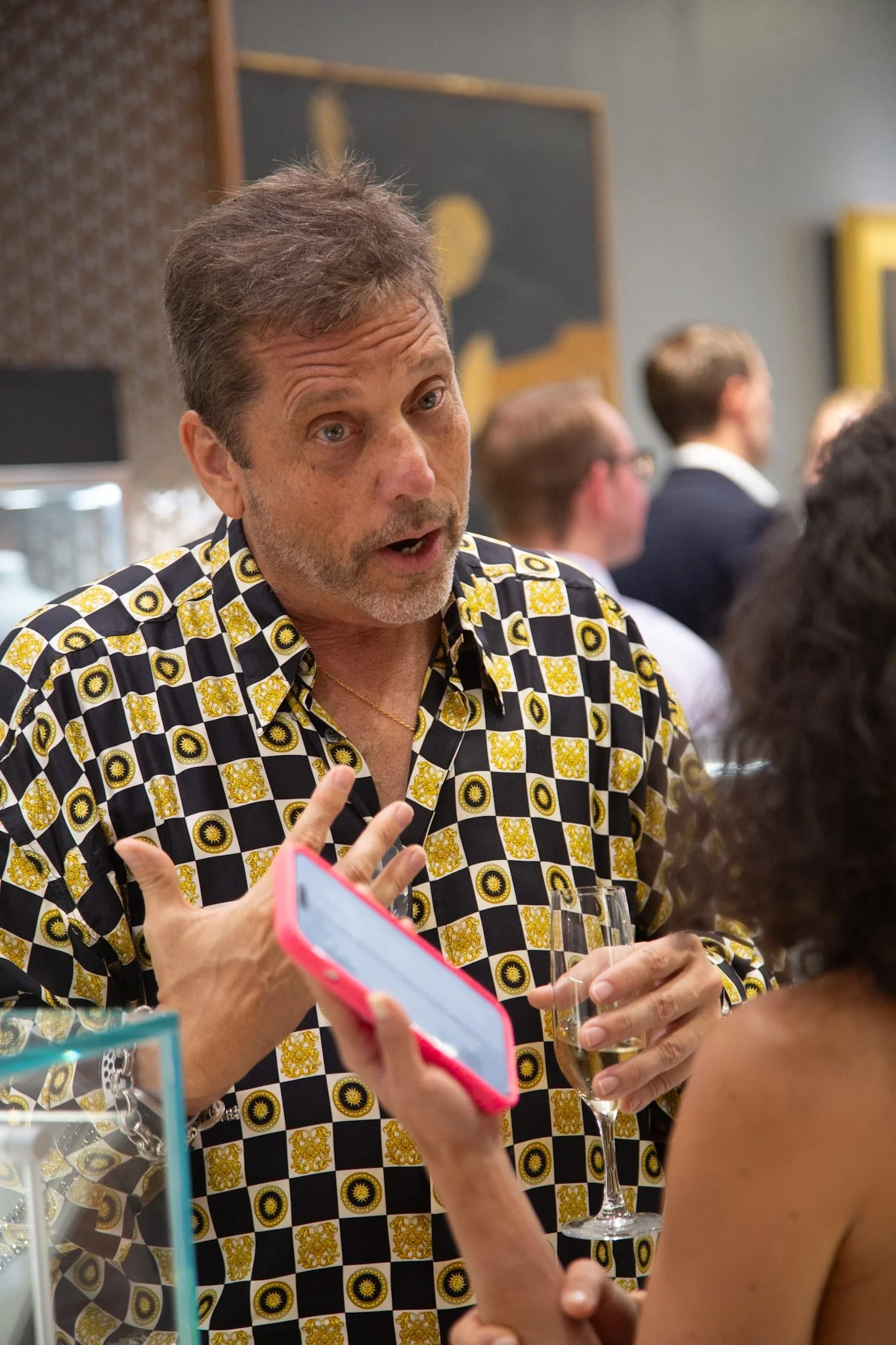 A middle-aged man with gray hair and beard, wearing a black and yellow patterned shirt, is talking passionately while holding a glass of wine at a social event. Several other people are in the background.