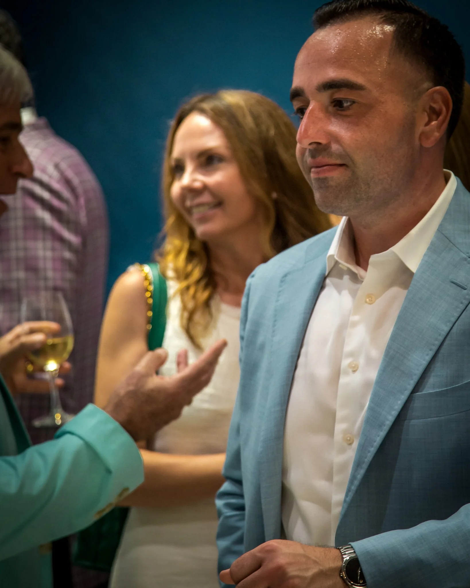 People in a social setting, with one man in a light blue blazer and white shirt in the foreground, engaging in conversation, and others talking and holding drinks in the background.