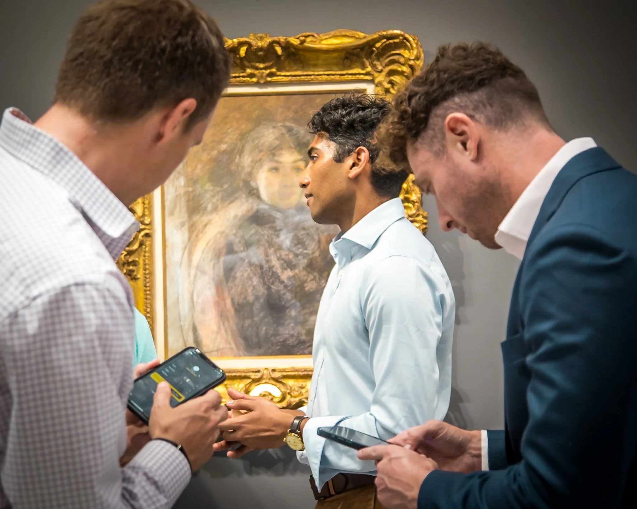 Three men standing in front of a painting, looking at their phones, with a woman partially visible in the background.