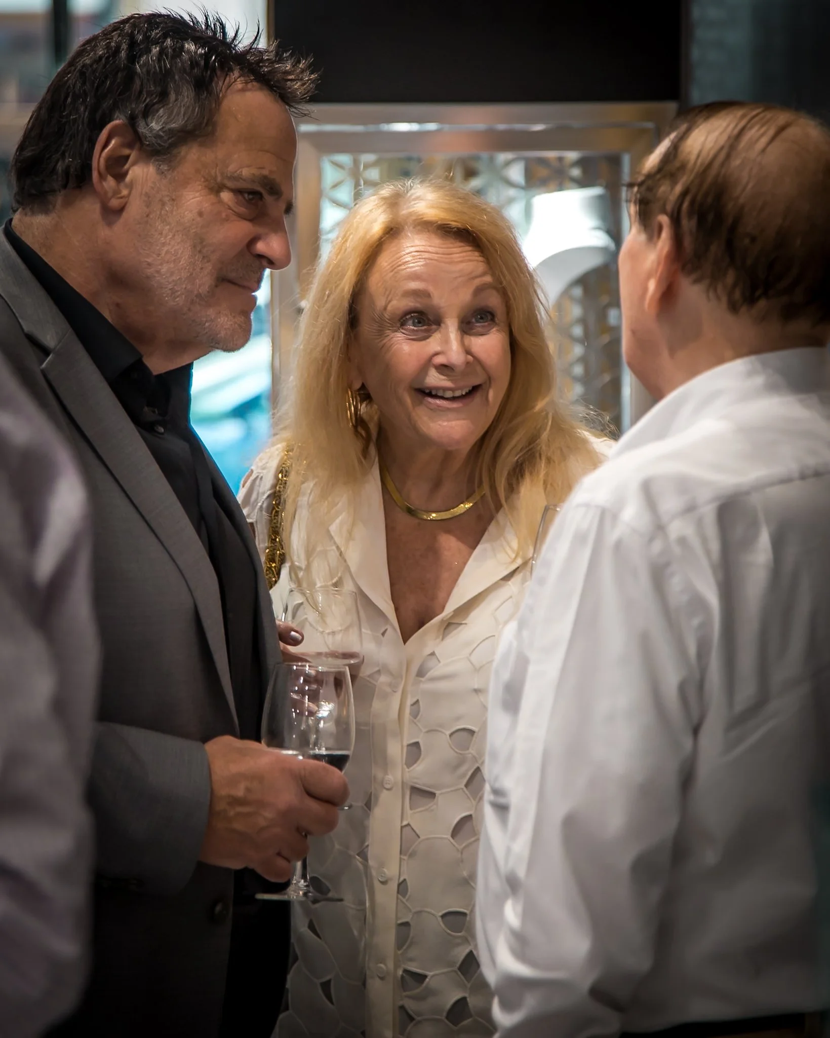 Three people having a conversation at a social event, one man holding a glass of red wine, a woman smiling, and a man with glasses turned away from the camera.