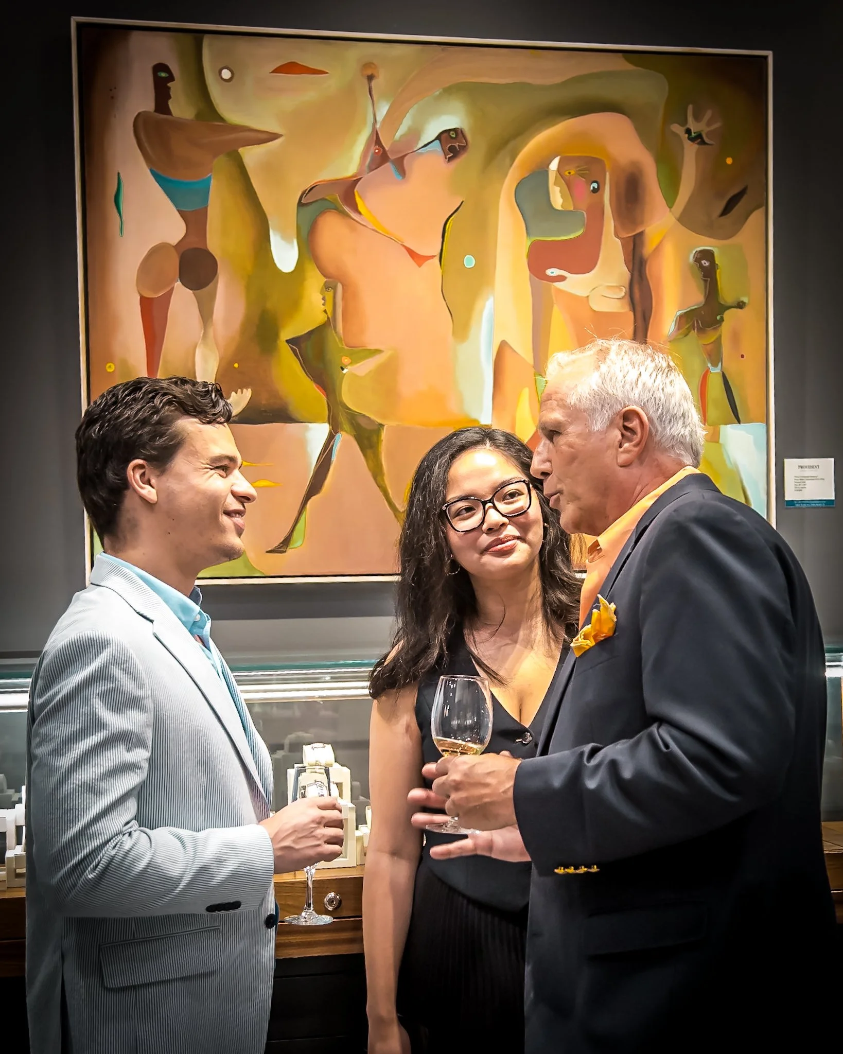 Three people in formal attire holding glasses of wine are having a conversation at a social event in an art gallery, with an abstract painting in the background.