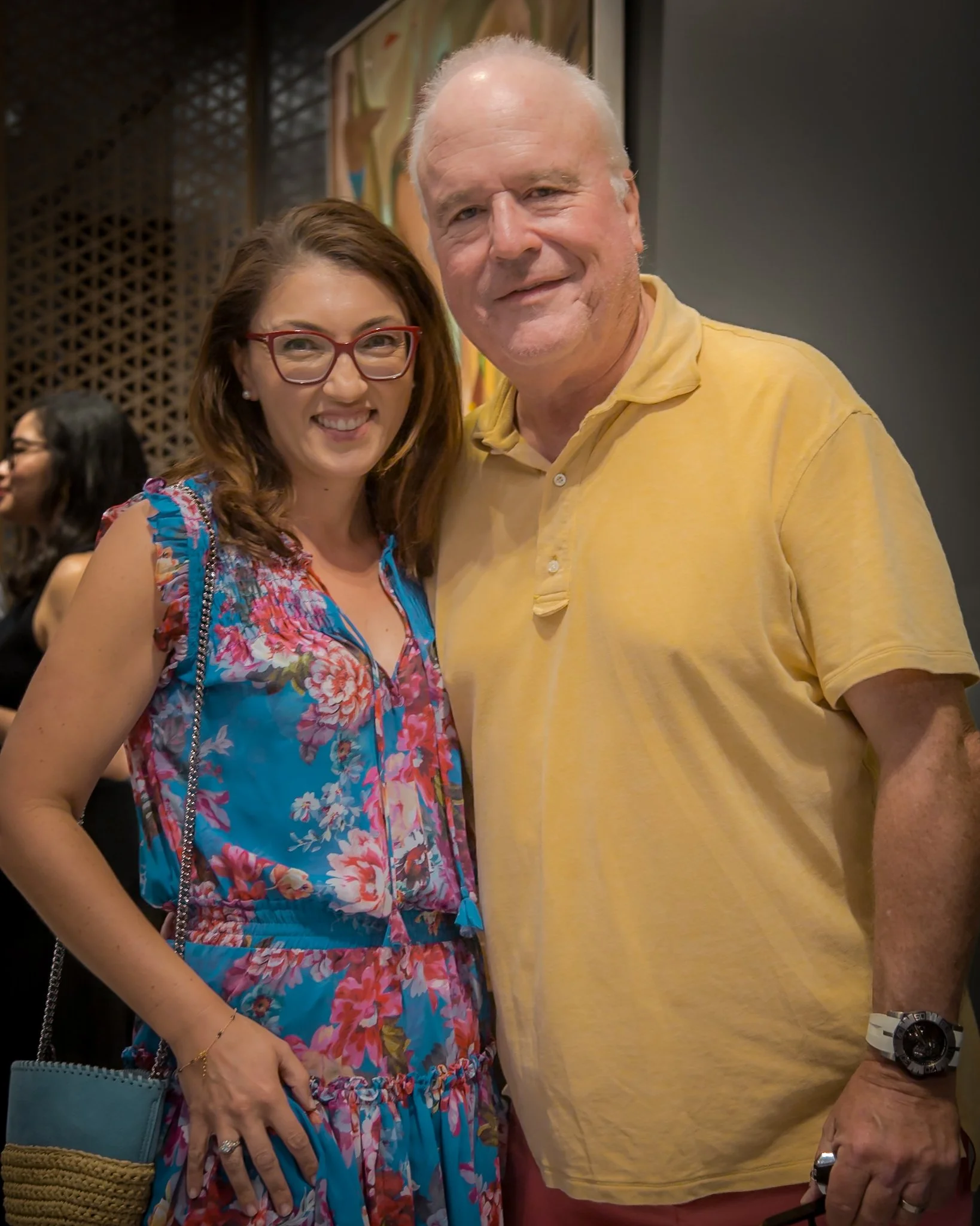 A woman in a colorful floral dress with red glasses and a woman in a yellow shirt standing close together and smiling at an indoor event.