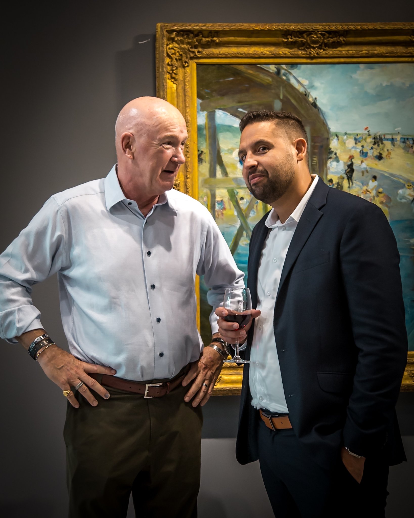 Two men in business attire at an art gallery, with a painting of a pier and beach scene in the background. One man is holding a glass of red wine.
