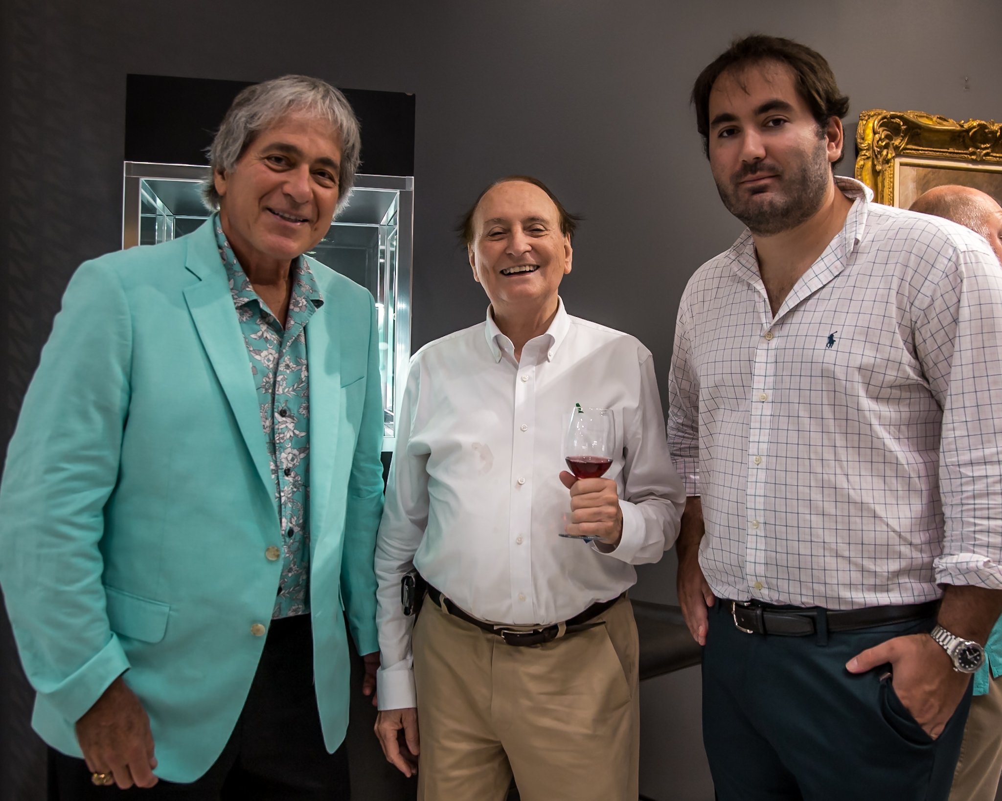 Three men standing together, smiling, one holding a glass of red wine, in an indoor setting with a dark wall and a framed artwork on the wall behind them.