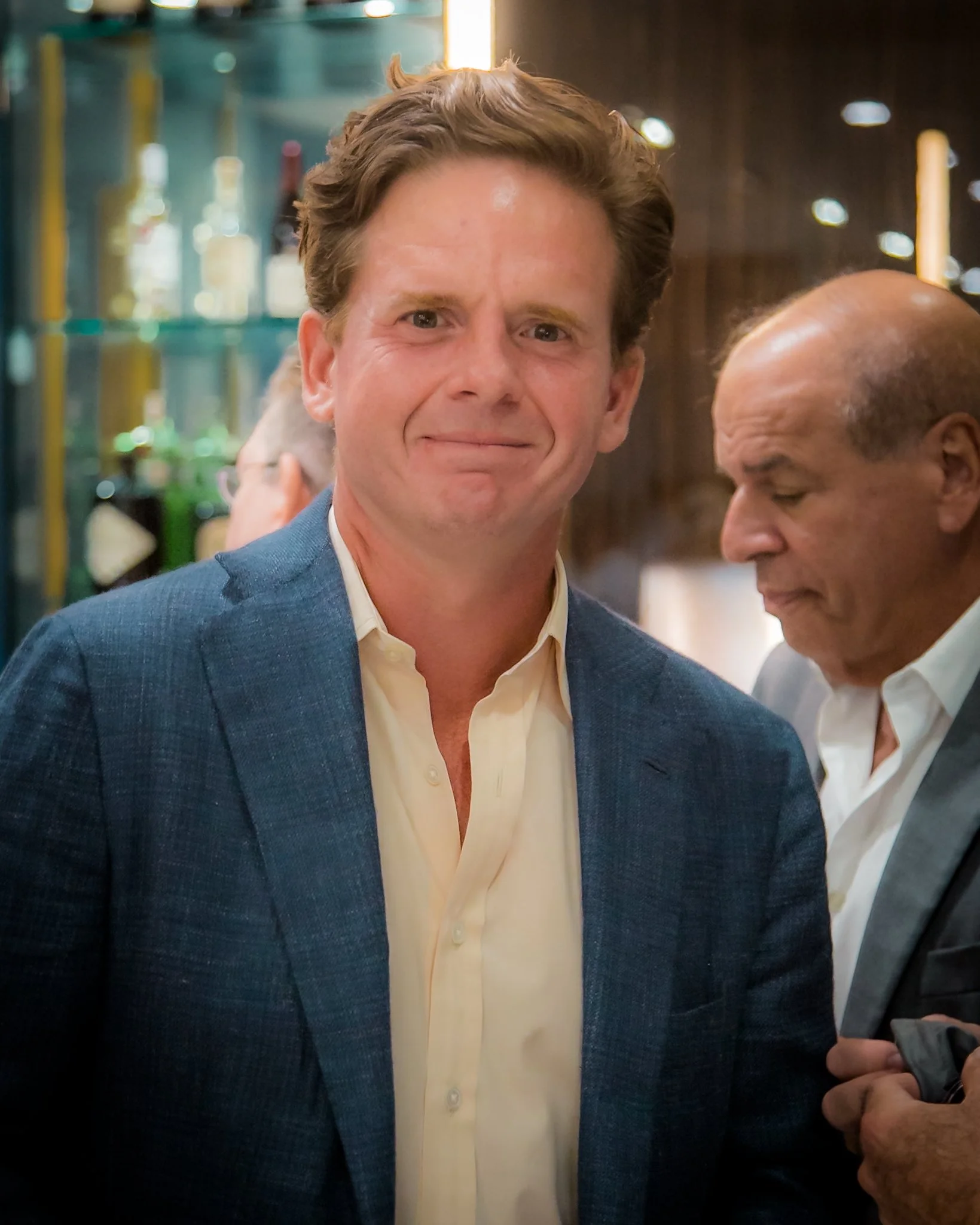 A man with brown hair, wearing a yellow shirt and blue blazer, faces the camera at a social event with blurred background.