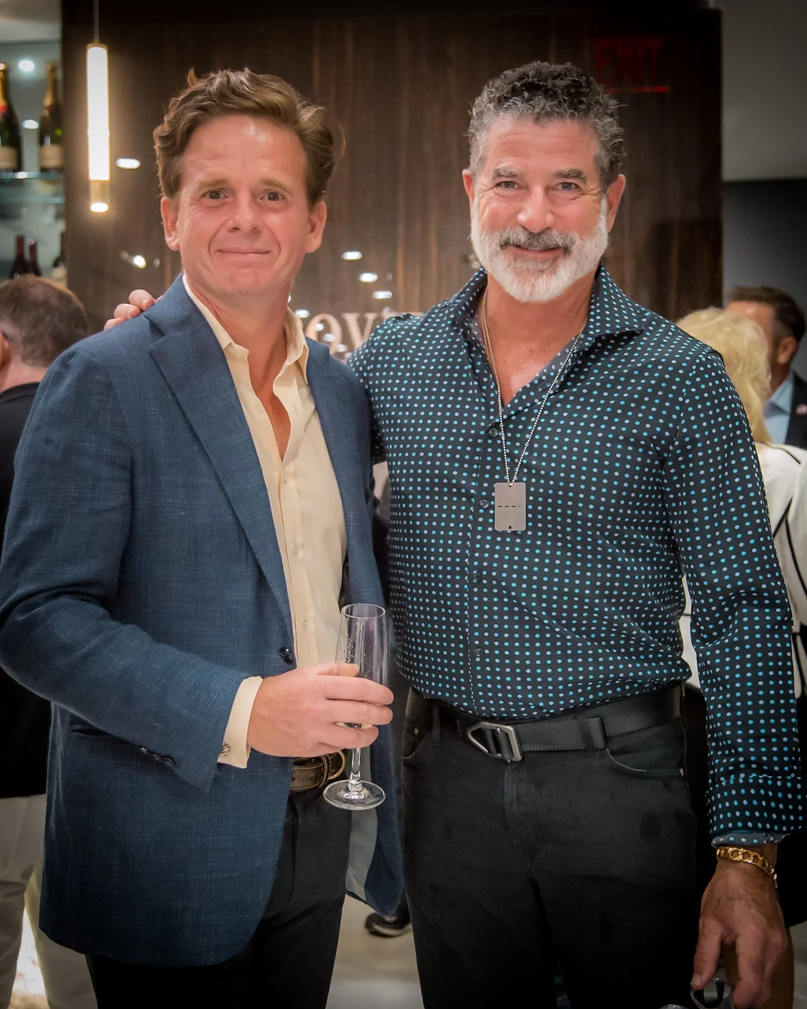 Two men at a social gathering posing for a photo. The man on the left is wearing a blue blazer and holding a glass of wine. The man on the right has gray hair and a beard, wearing a black shirt with small blue dots and a dog tag necklace.