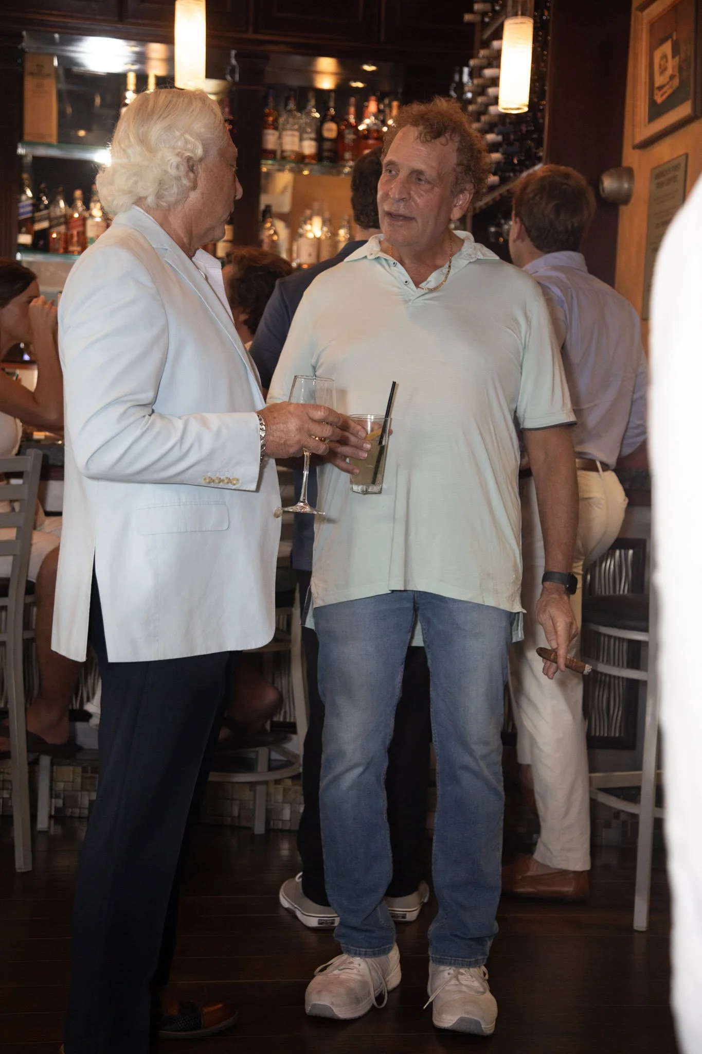 Two men are engaged in conversation at a bar or restaurant. The man on the left has white hair and is wearing a white blazer and dark pants, holding a glass of champagne. The man on the right has curly hair, is wearing a light-colored polo shirt, jea