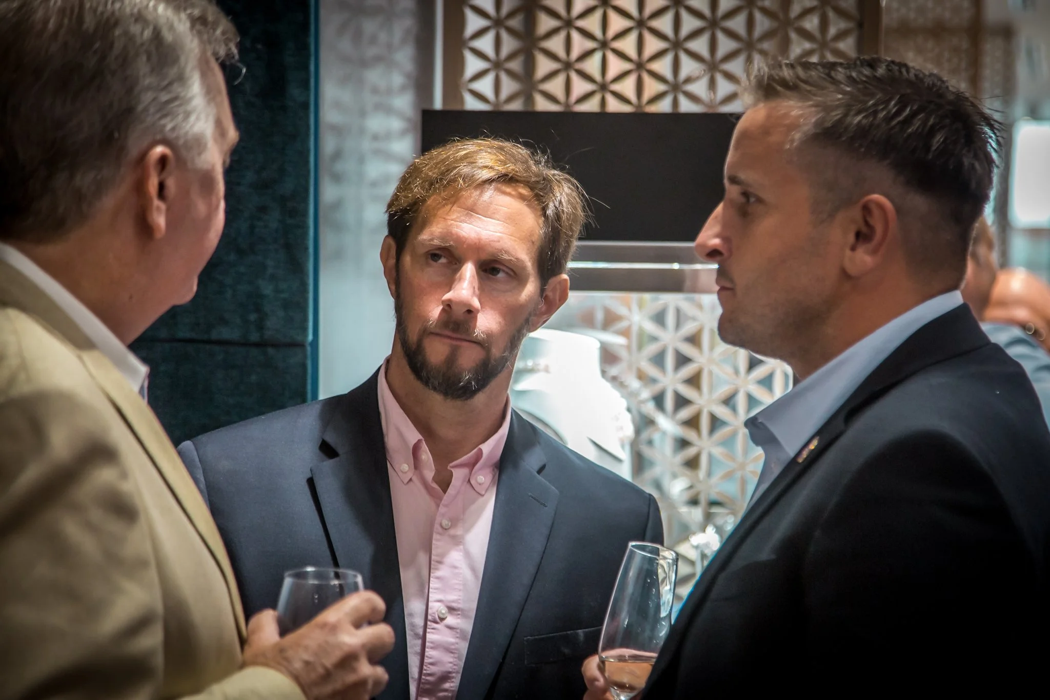 Three men in suits engaging in a conversation at a social event, two holding glasses of wine.
