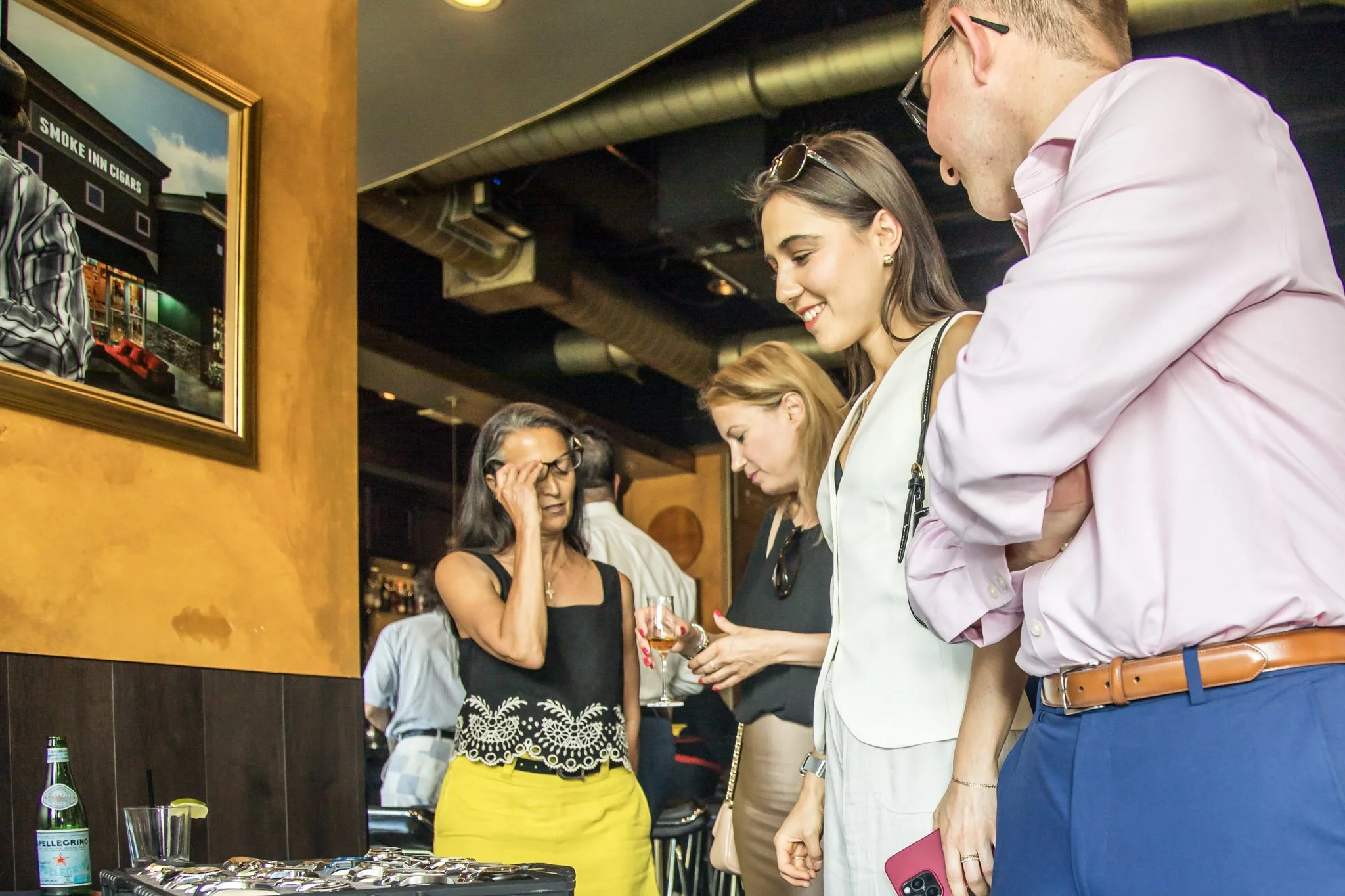 Four people standing in a group inside a bar or restaurant, looking at jewelry or small items. The woman on the left is wearing a black sleeveless top with lace and yellow pants. The woman has a wine glass and is holding a jewelry box. The man on the