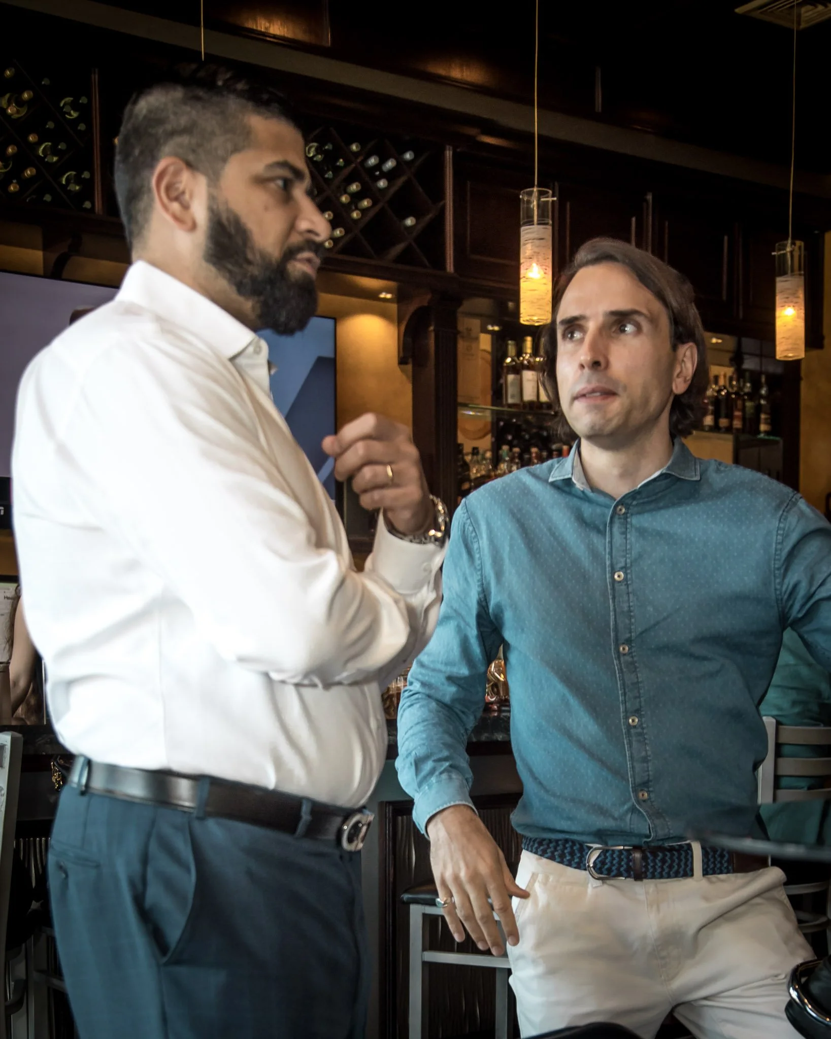 Two men engaged in conversation at a bar, one with a beard in a white shirt, and the other with wavy hair in a blue shirt.