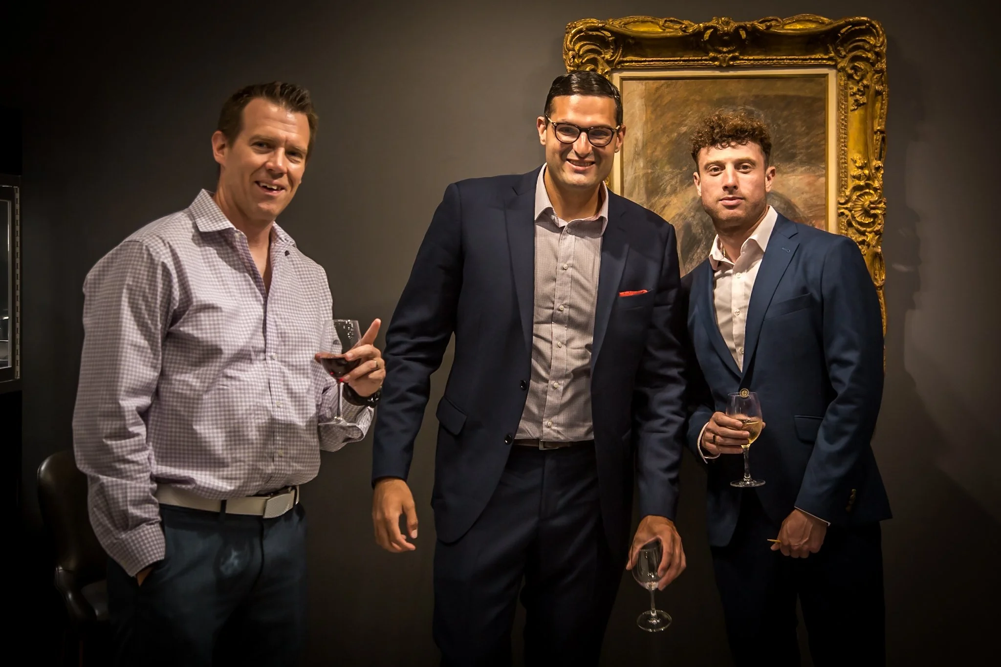 Three men dressed in suits at a social gathering, holding wine glasses and smiling, standing in front of a dark wall and a framed painting.