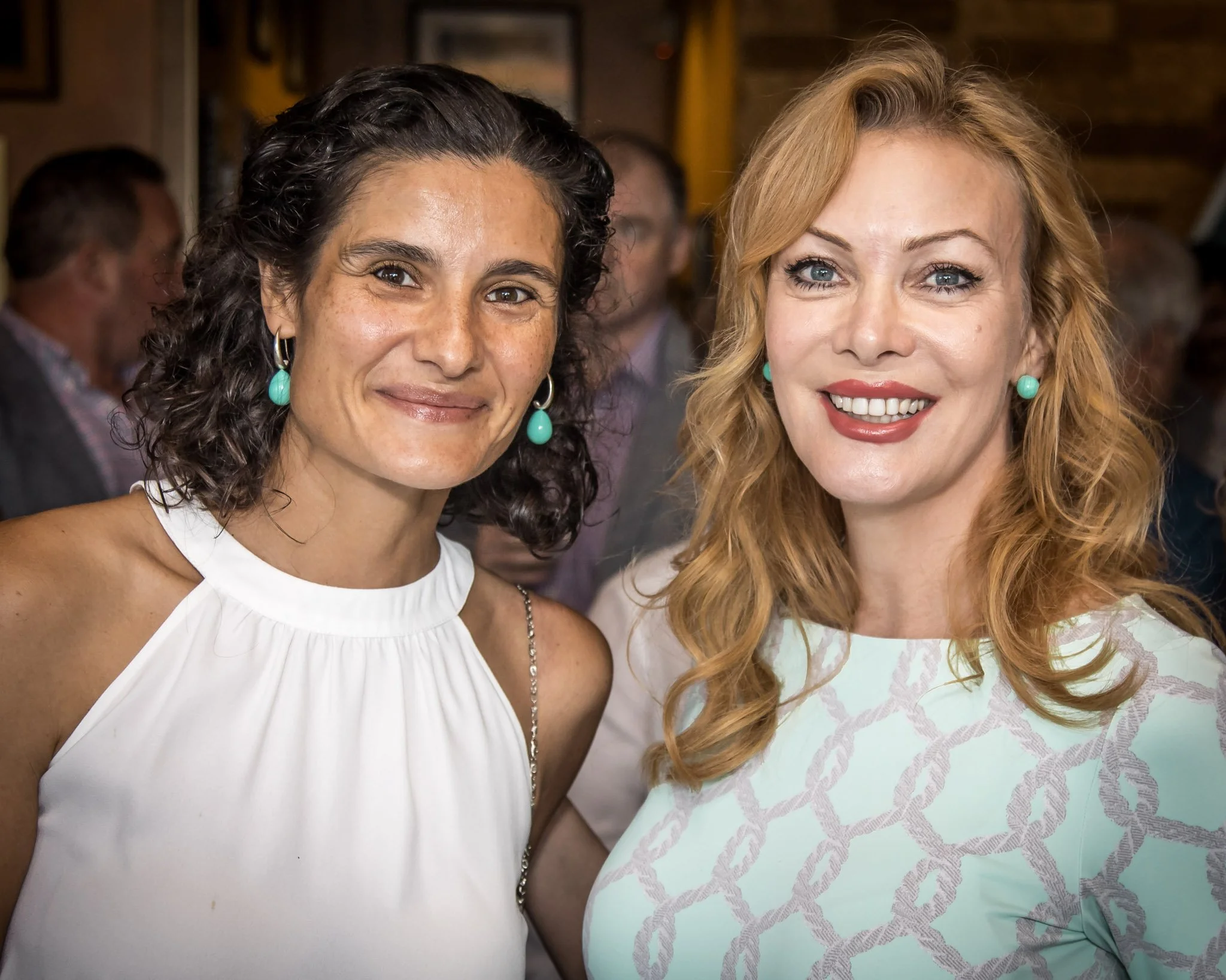 Two women smiling at a social gathering, one with dark curly hair wearing a white sleeveless top and turquoise earrings, the other with red hair wearing a patterned light green dress and matching earrings, with blurred people in the background.
