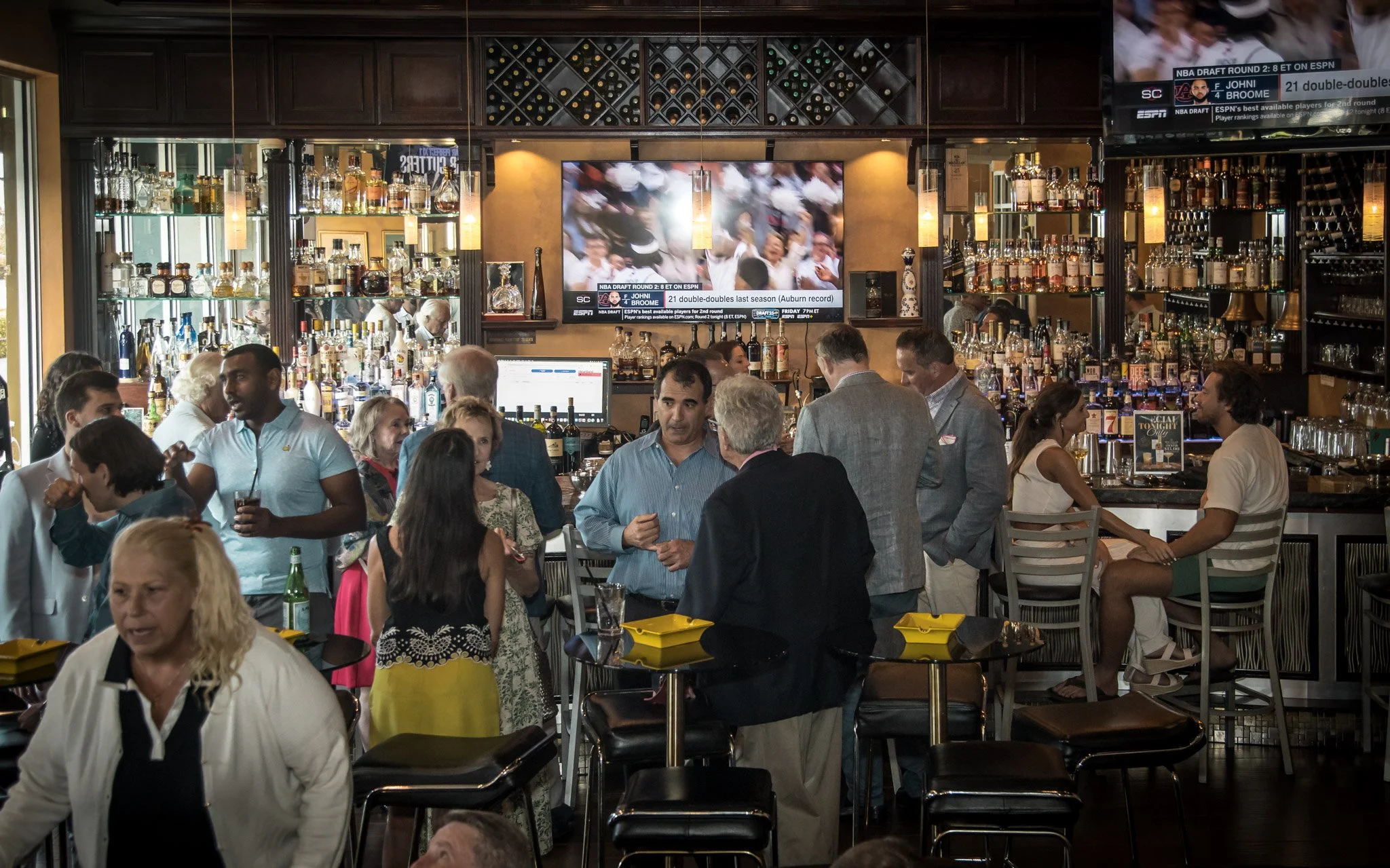 A busy bar scene with people socializing, drinks on the tables, and a large TV screen displaying a sports event in the background.